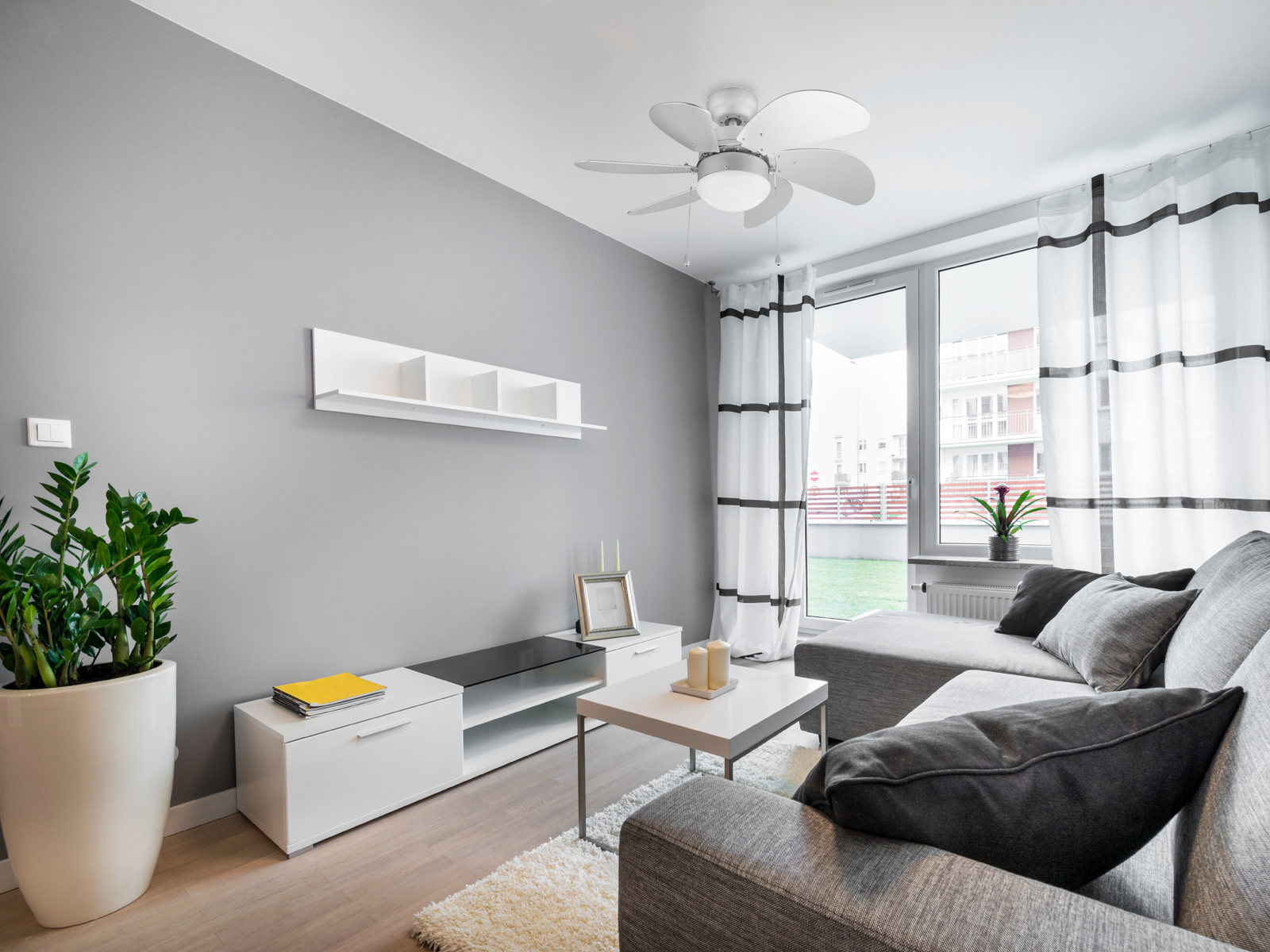 Wohnzimmer mit grauen Wänden und Möbeln, weißen Vorhängen, Deckenventilator und grüner Pflanze.