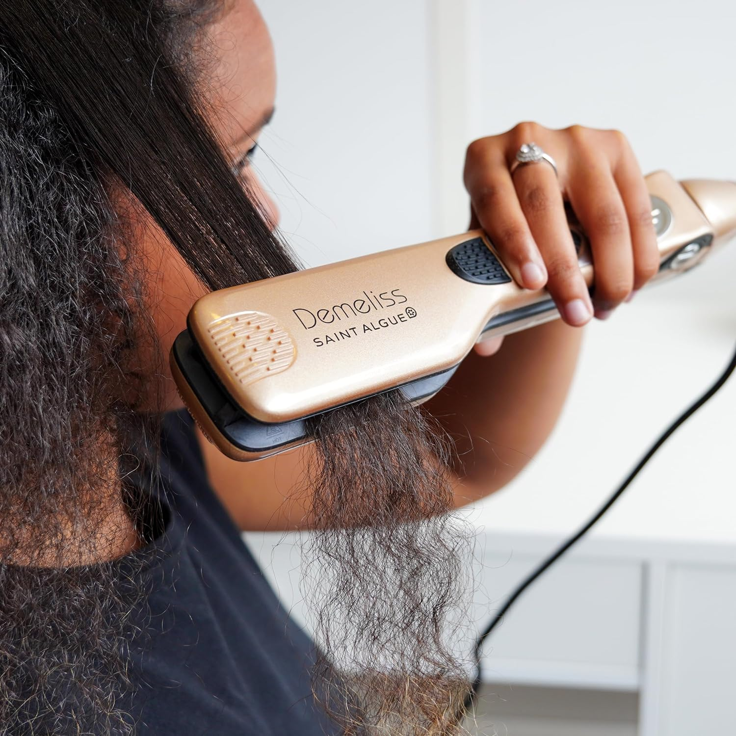 Eine Person, die einen goldenen Haarglätter auf dunklem, welligem Haar verwendet, mit weißem Hintergrund.