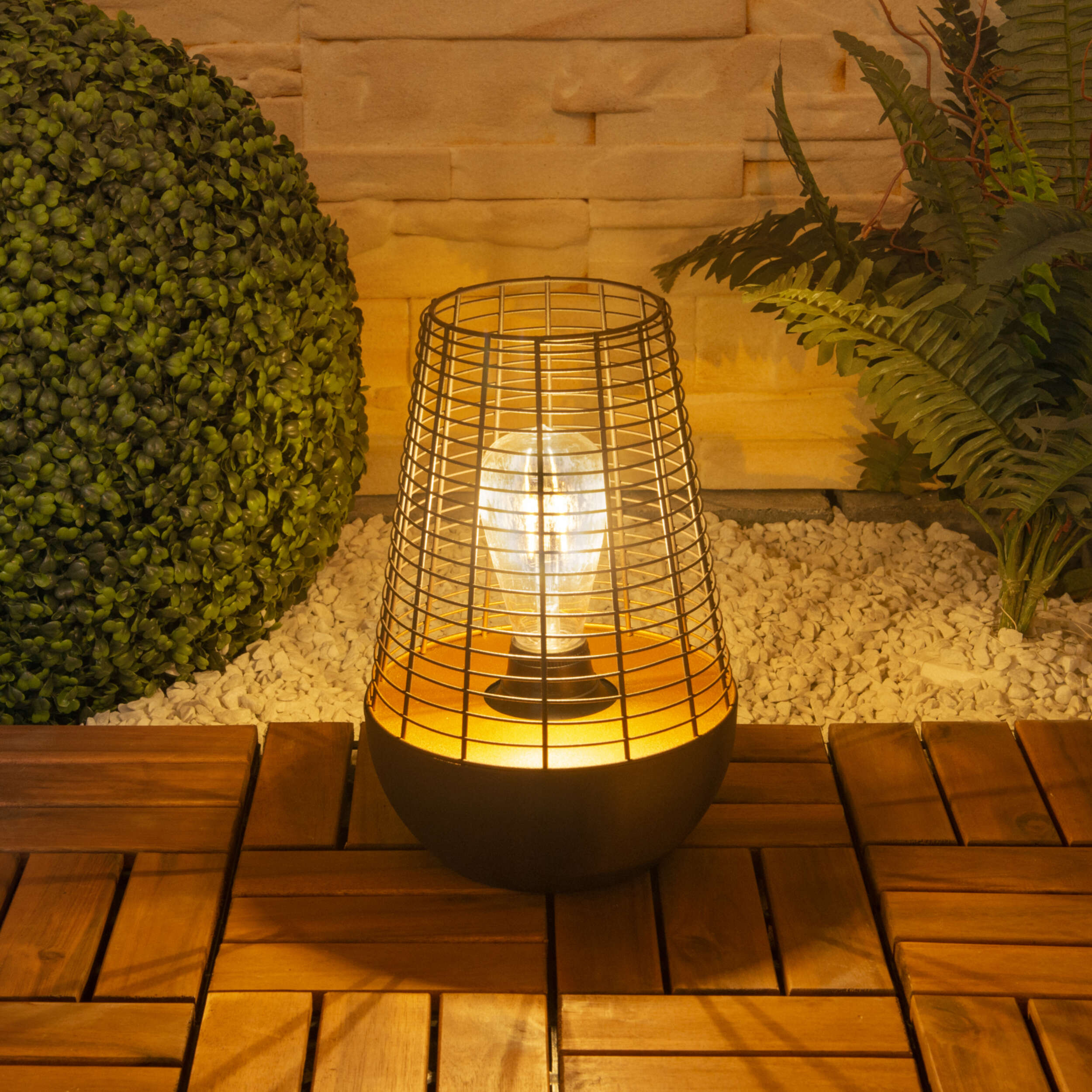 Außenlampe mit Drahtkäfig um eine Glühbirne, auf einem Holzdeck mit Grünpflanzen.