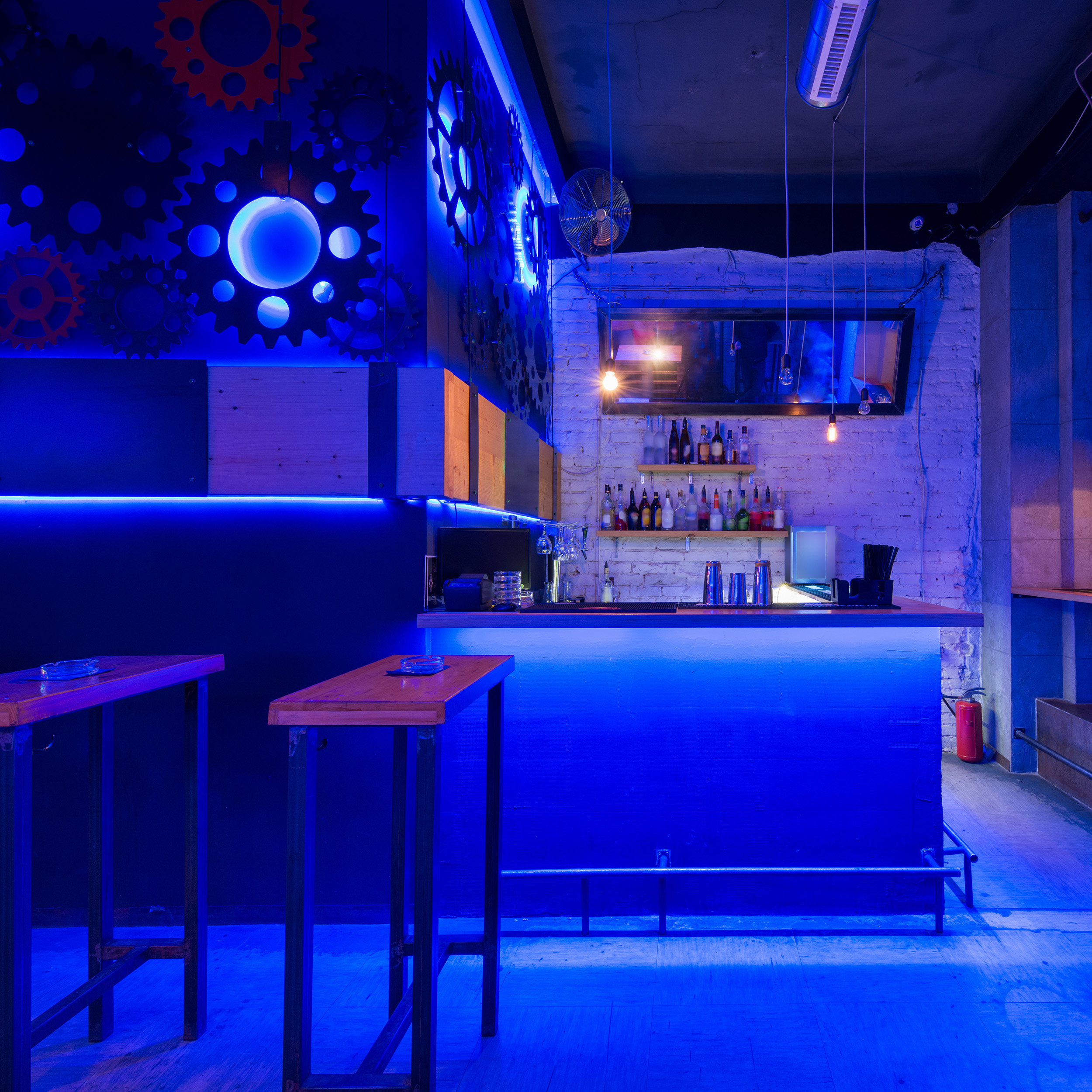 Innenansicht einer Bar mit blauen Neonlichtern, Tischen und Flaschen auf Regalen.