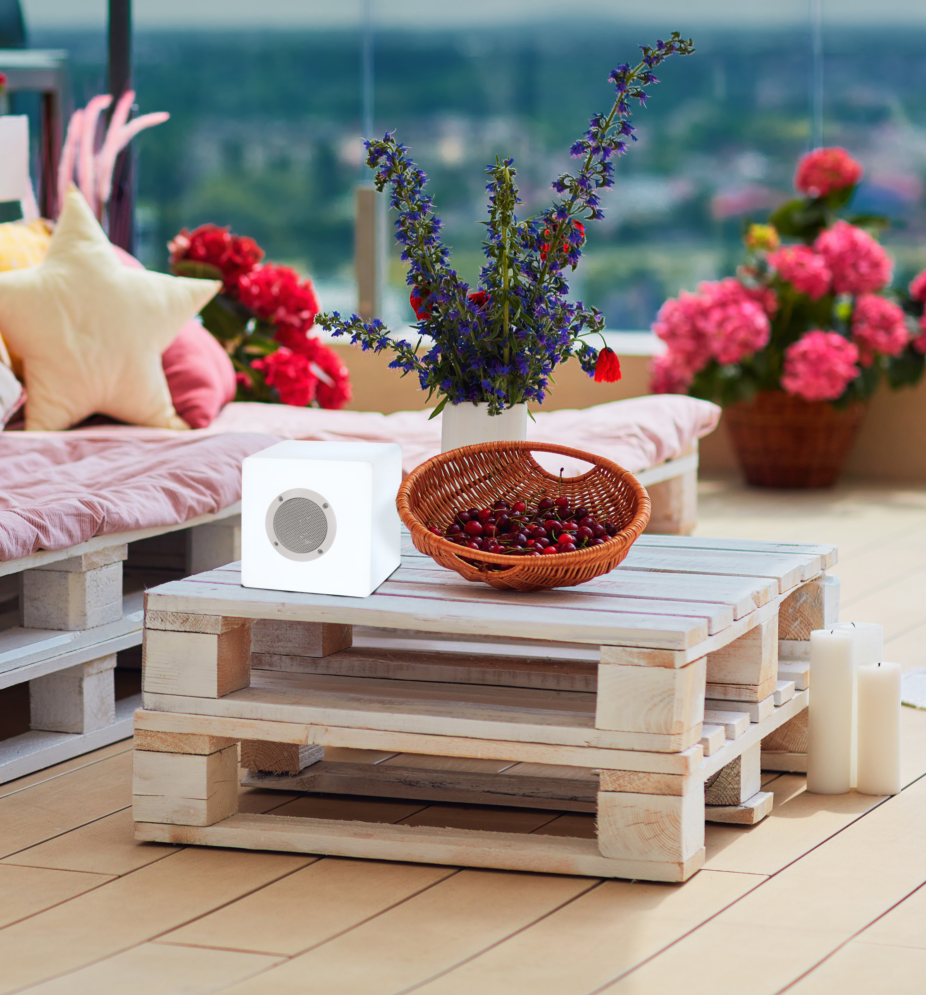 Szene im Freien mit weißem Tisch, Blumen, Kerzen und einem Lautsprecher auf einem Balkon.