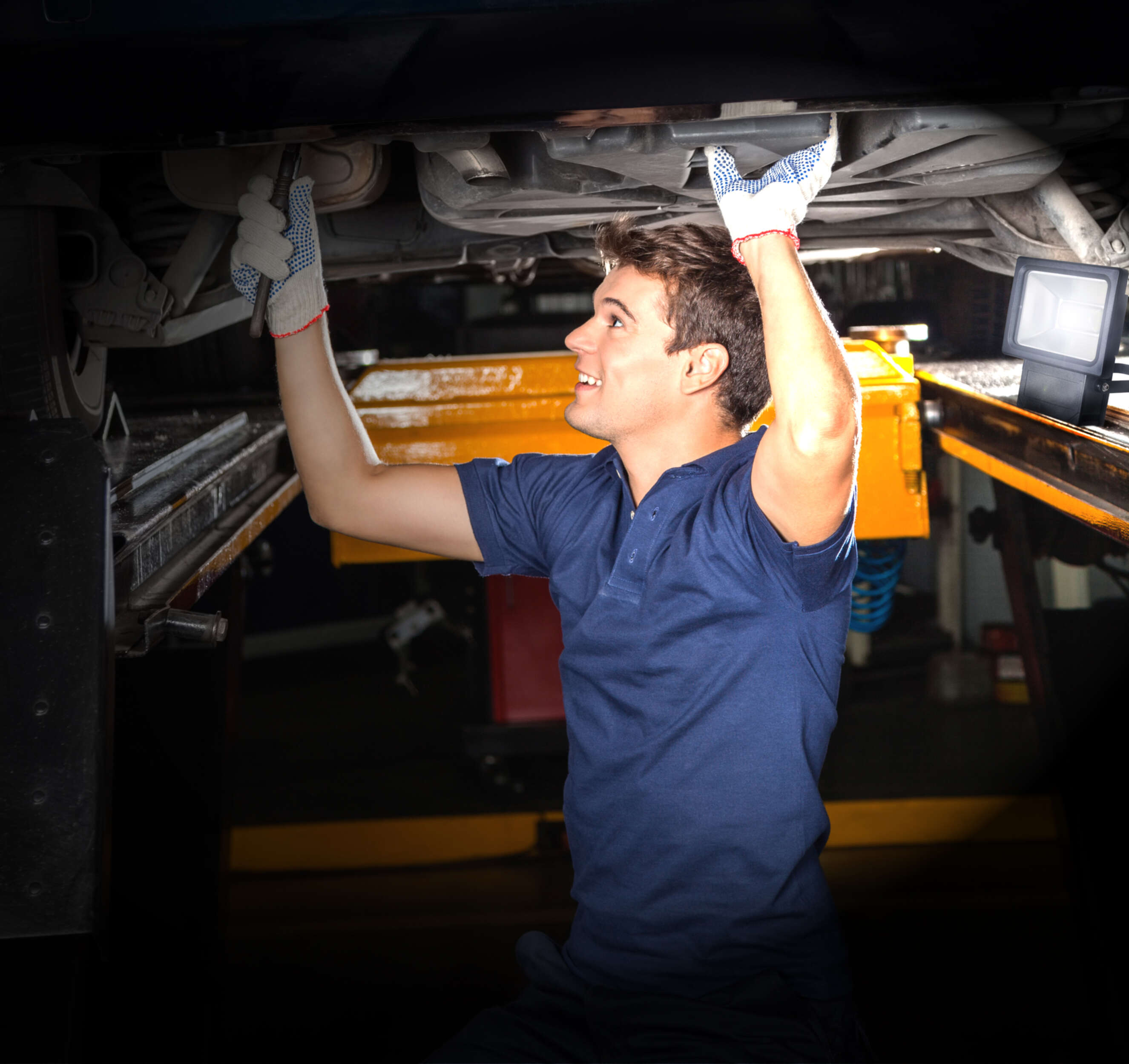 Ein Mann arbeitet von unten an einem Auto, trägt Handschuhe, mit einer leuchtenden Lampe in der Nähe.