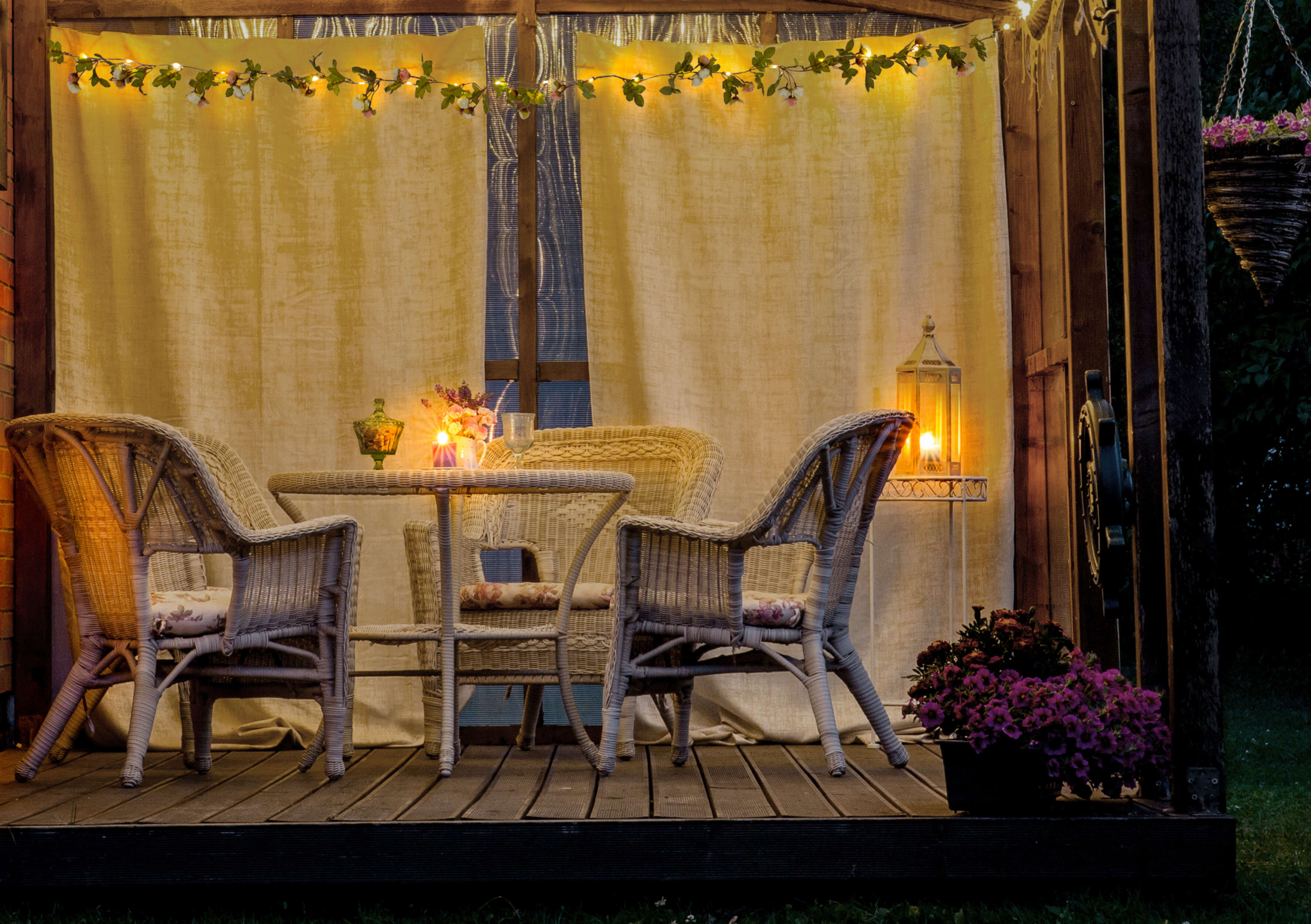 Eine Holzterrasse mit Stühlen, einem Tisch und einer Hängelampe. Lichterketten und Vorhänge sind vorhanden.