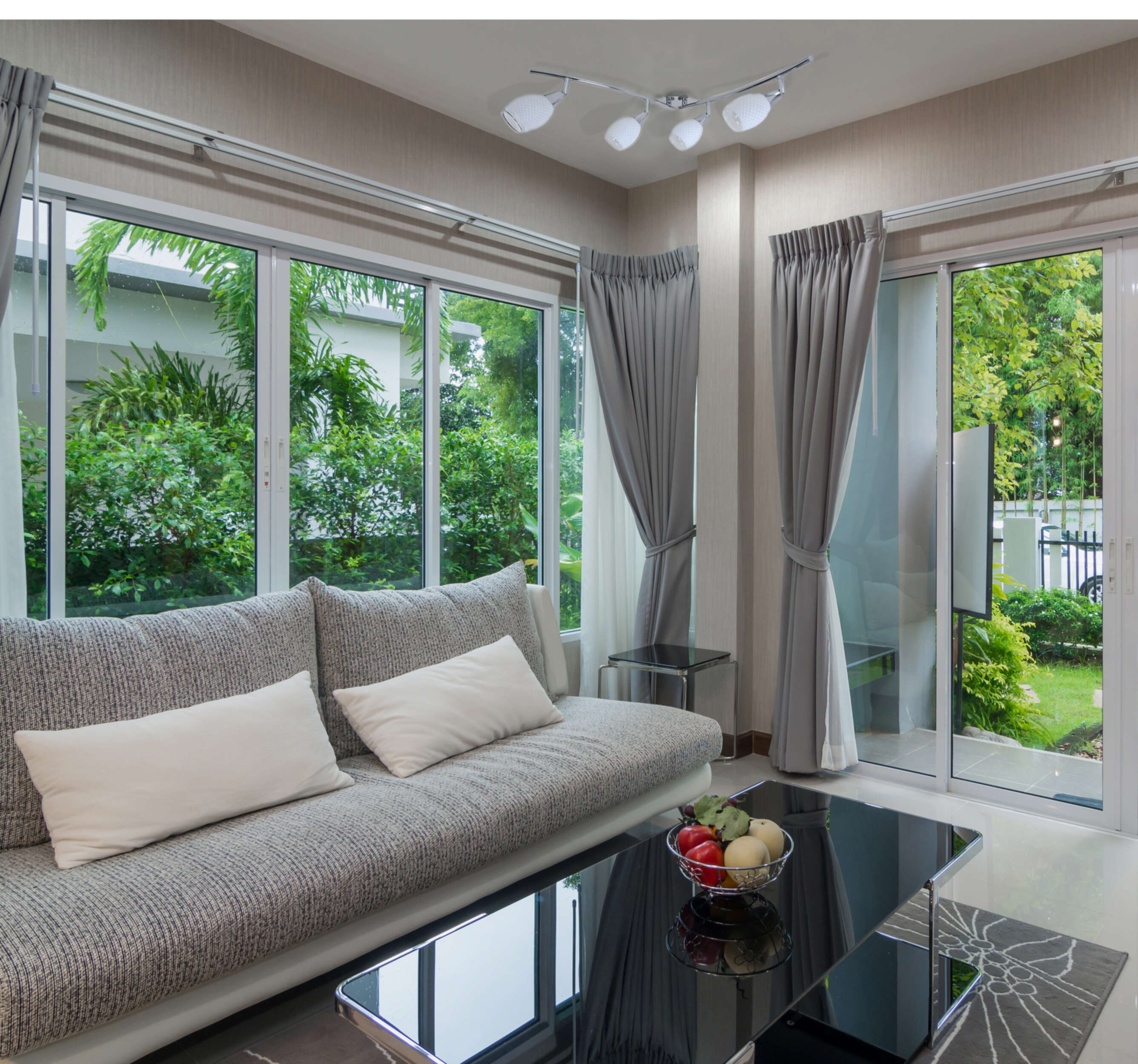 Wohnzimmerinterieur mit grauem Sofa, Fenstern, Vorhängen, Tisch mit Obst und Blick nach draußen.
