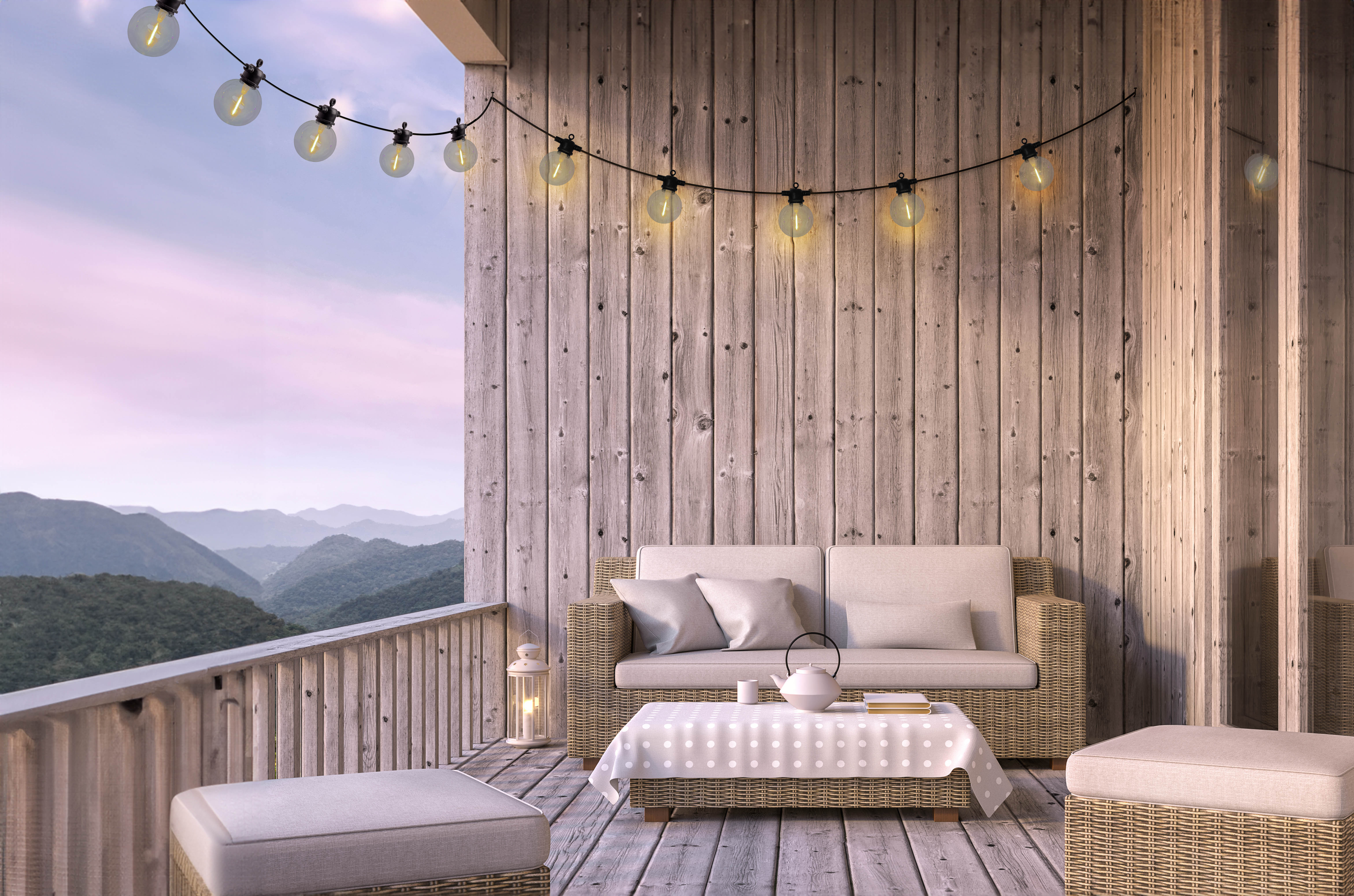 Holzveranda mit Korbsofa, Tisch, Lichtern und Blick auf die Berge.
