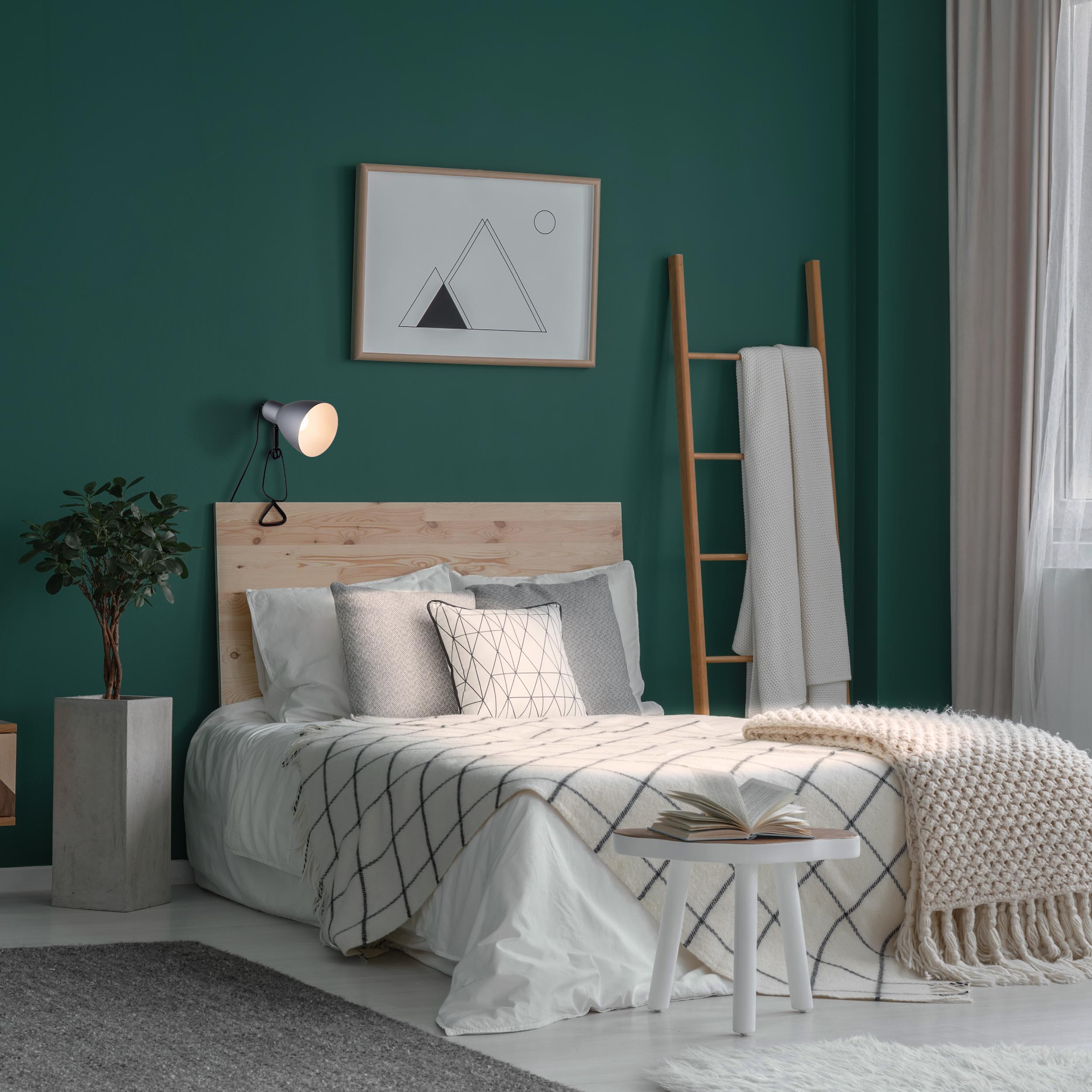 Schlafzimmer mit grüner Wand, Bett, Holz-Kopfteil, weißer Decke, Deko und einem kleinen Tisch mit Buch.