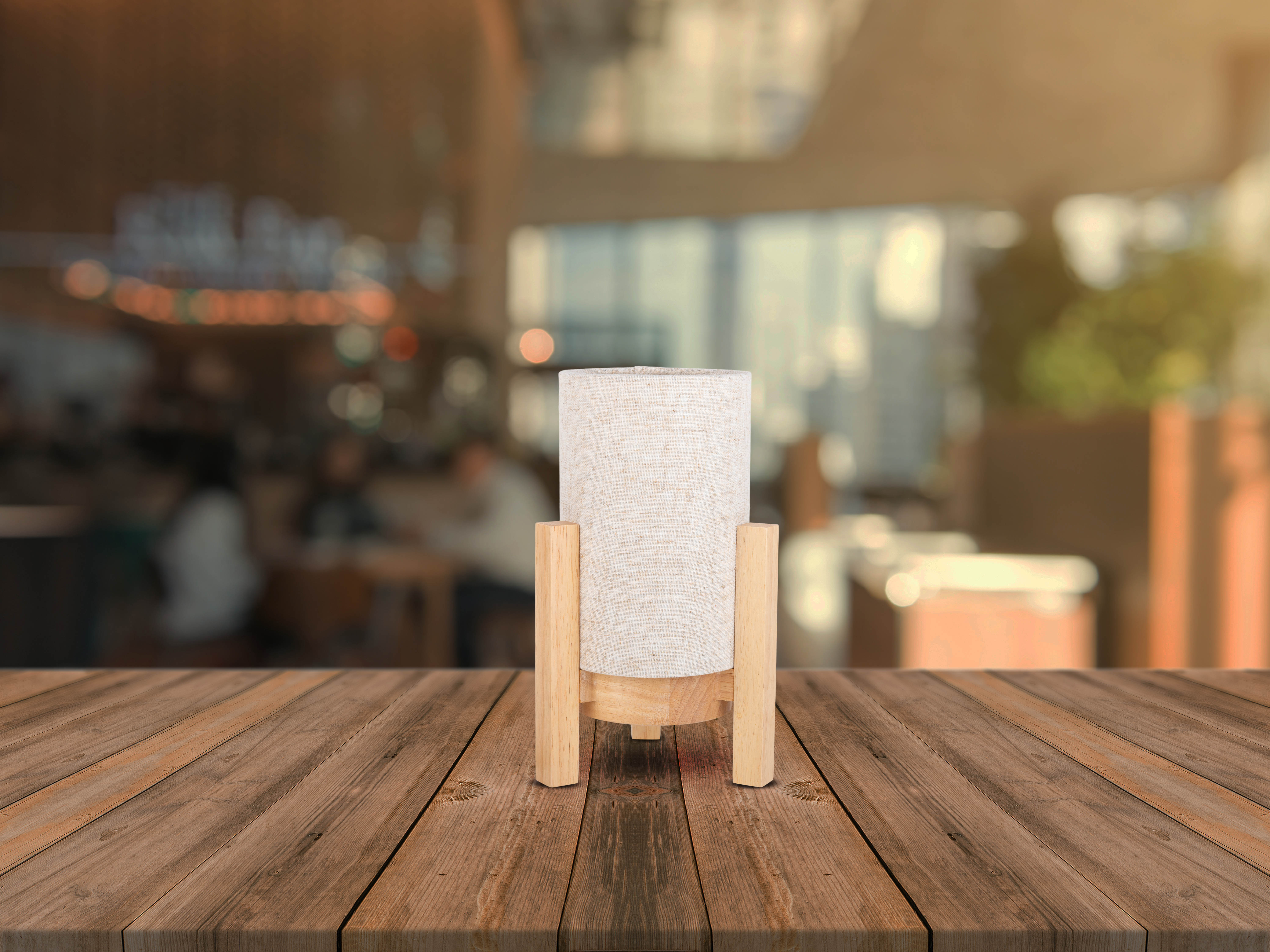 Eine Tischlampe mit zylindrischem Stoffschirm auf Holzsockel, auf einem Holztisch, mit unscharfem Hintergrund.