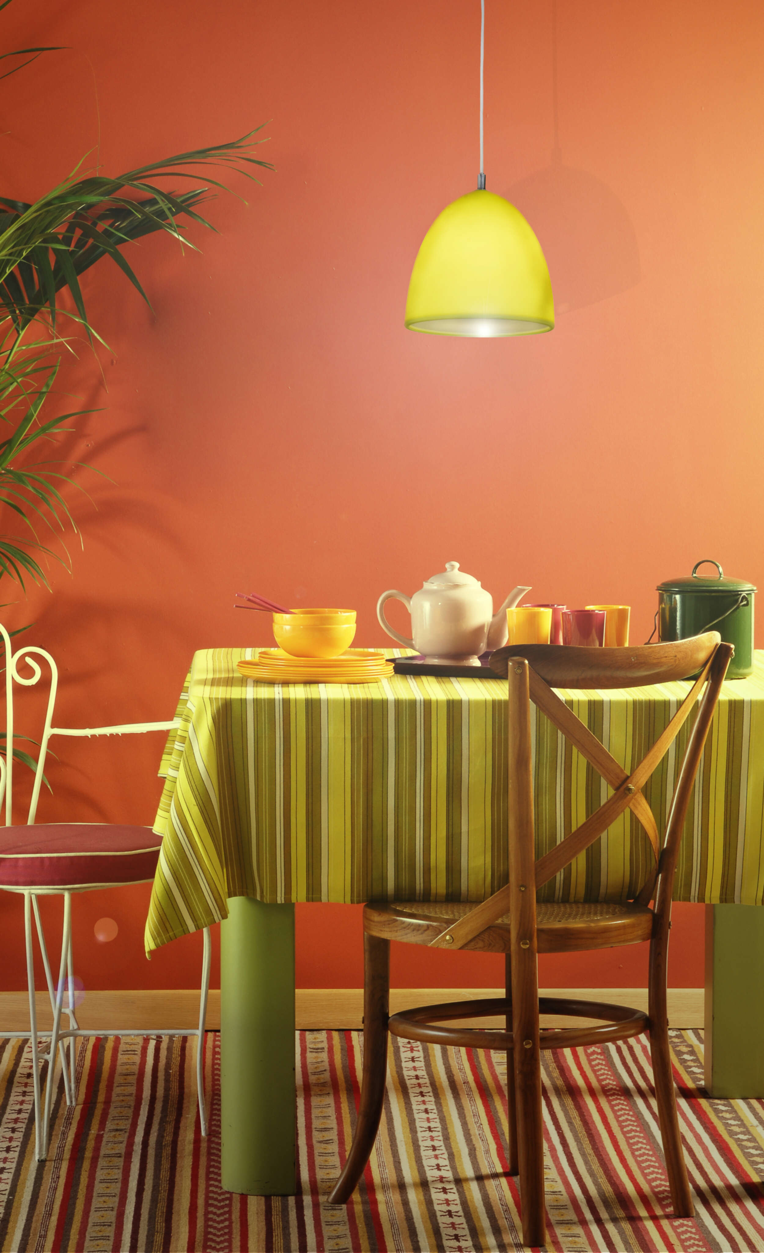 Ein Esstisch mit grün gestreifter Tischdecke, gelber Pendelleuchte und roter Wand.