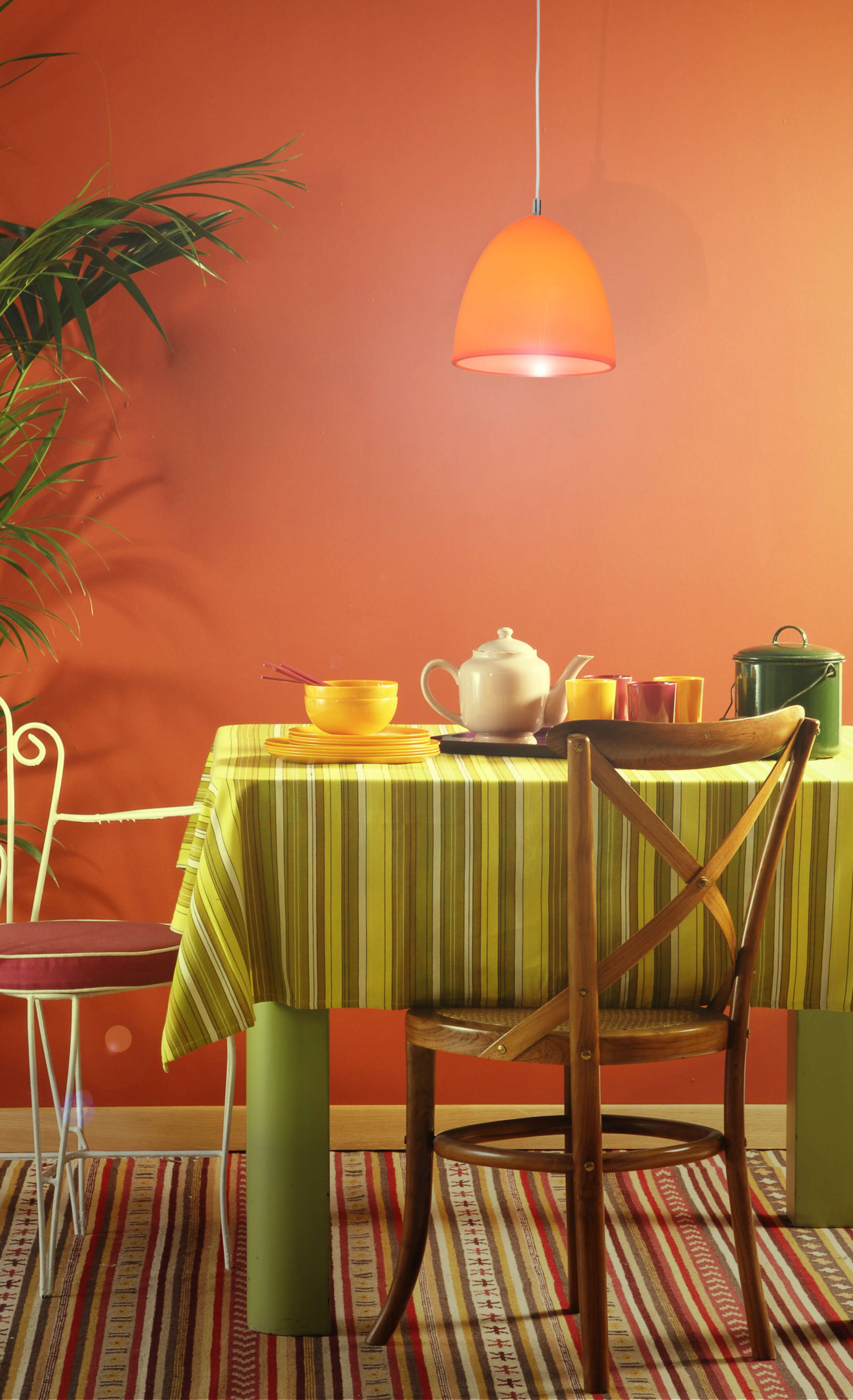 Ein Esstisch mit bunter Tischdecke, einer orangefarbenen Hängelampe und einer Pflanze. Die Wand ist orange.