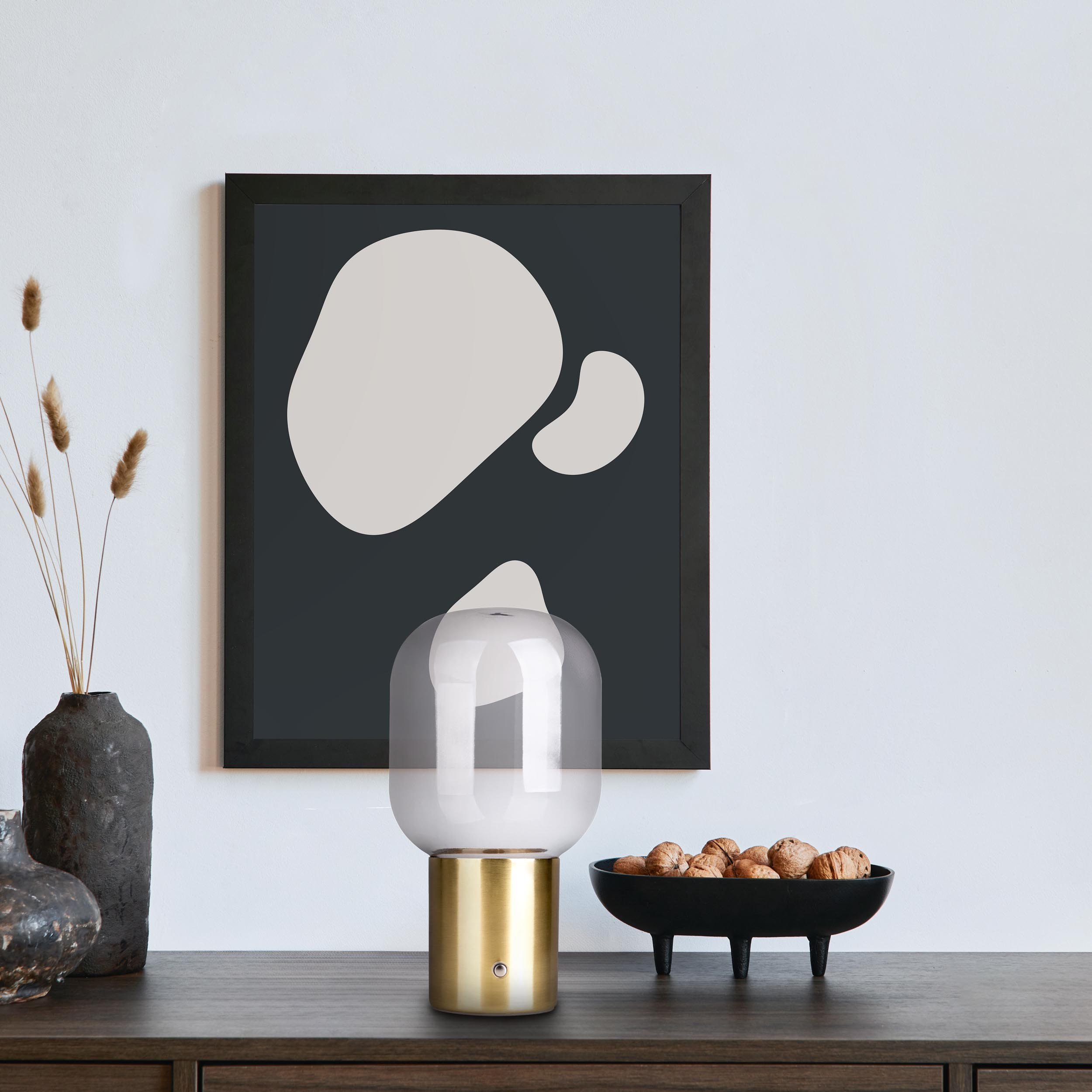 Ein gerahmtes Kunstwerk und eine Lampe stehen auf einem Holztisch. Eine Schüssel mit Walnüssen und eine Vase.