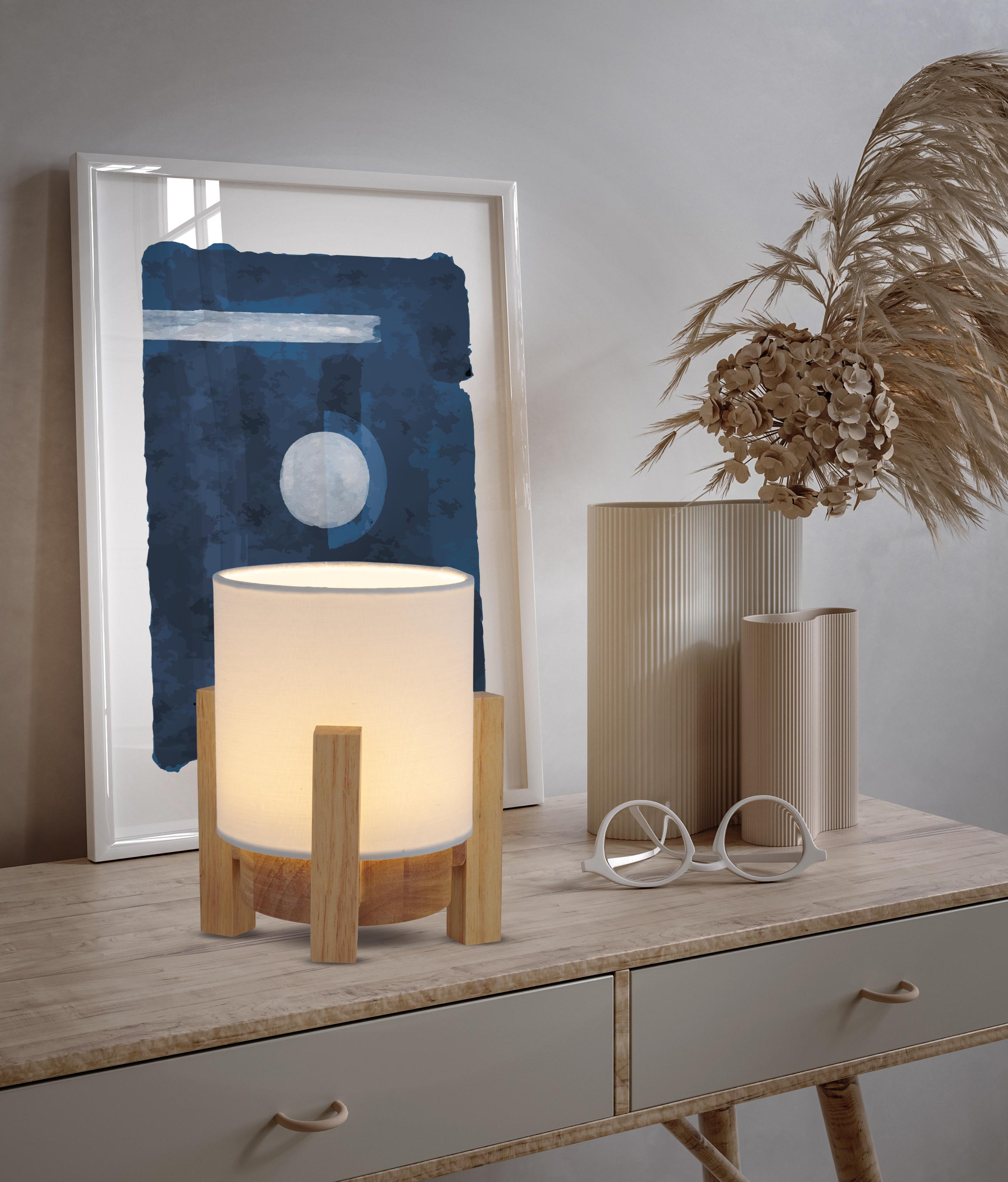Eine beleuchtete Lampe mit Holzsockel steht auf einem Tisch. Ein Gemälde, Vasen und eine Brille befinden sich ebenfalls auf dem Tisch.
