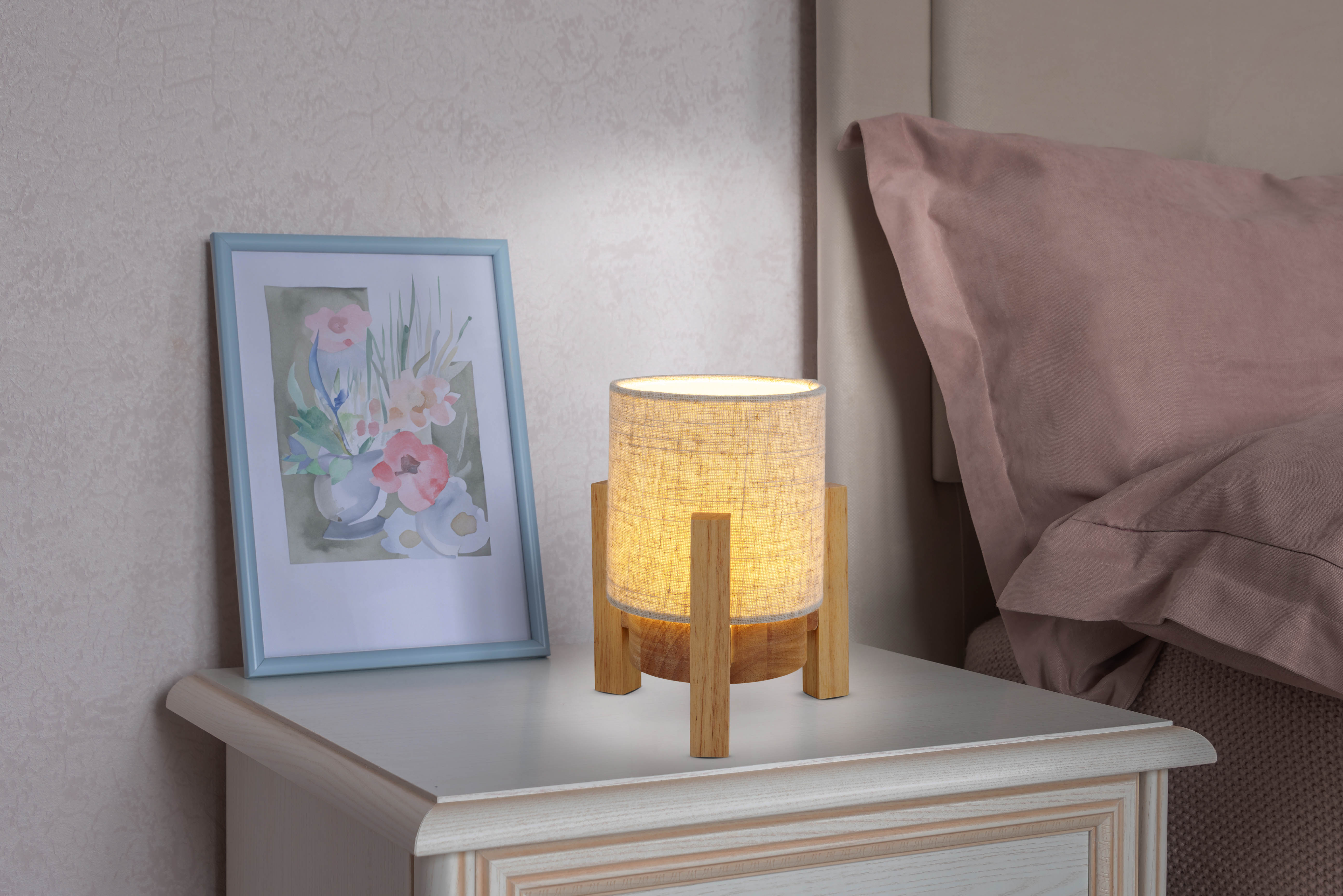 Lampe auf einem Nachttisch. Die Lampe hat einen beigen Schirm und einen Holzfuß. Bilderrahmen sichtbar.