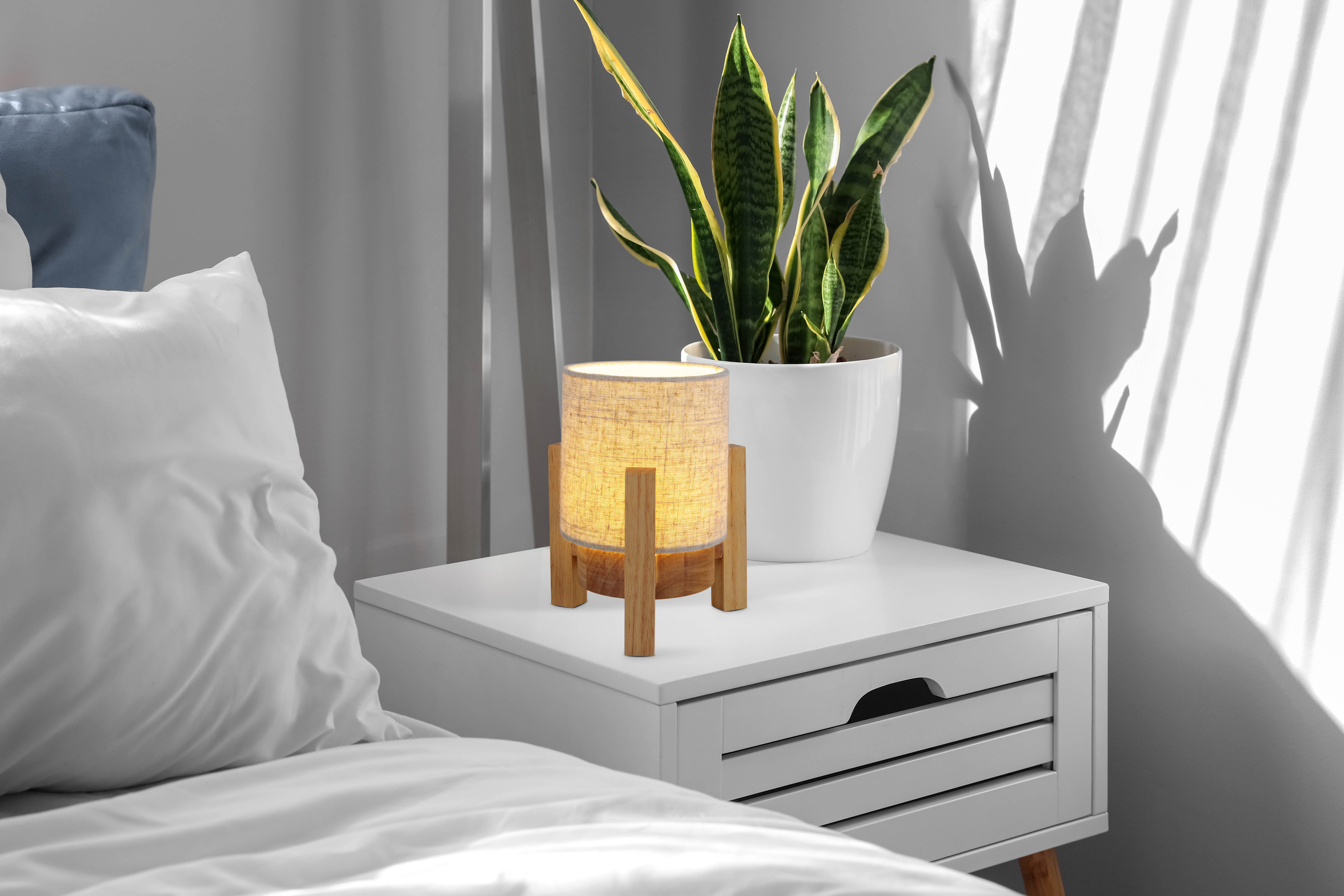 Eine Lampe auf einem Nachttisch. Die Lampe hat einen Holzsockel und einen Leinenschirm. Daneben eine Pflanze.