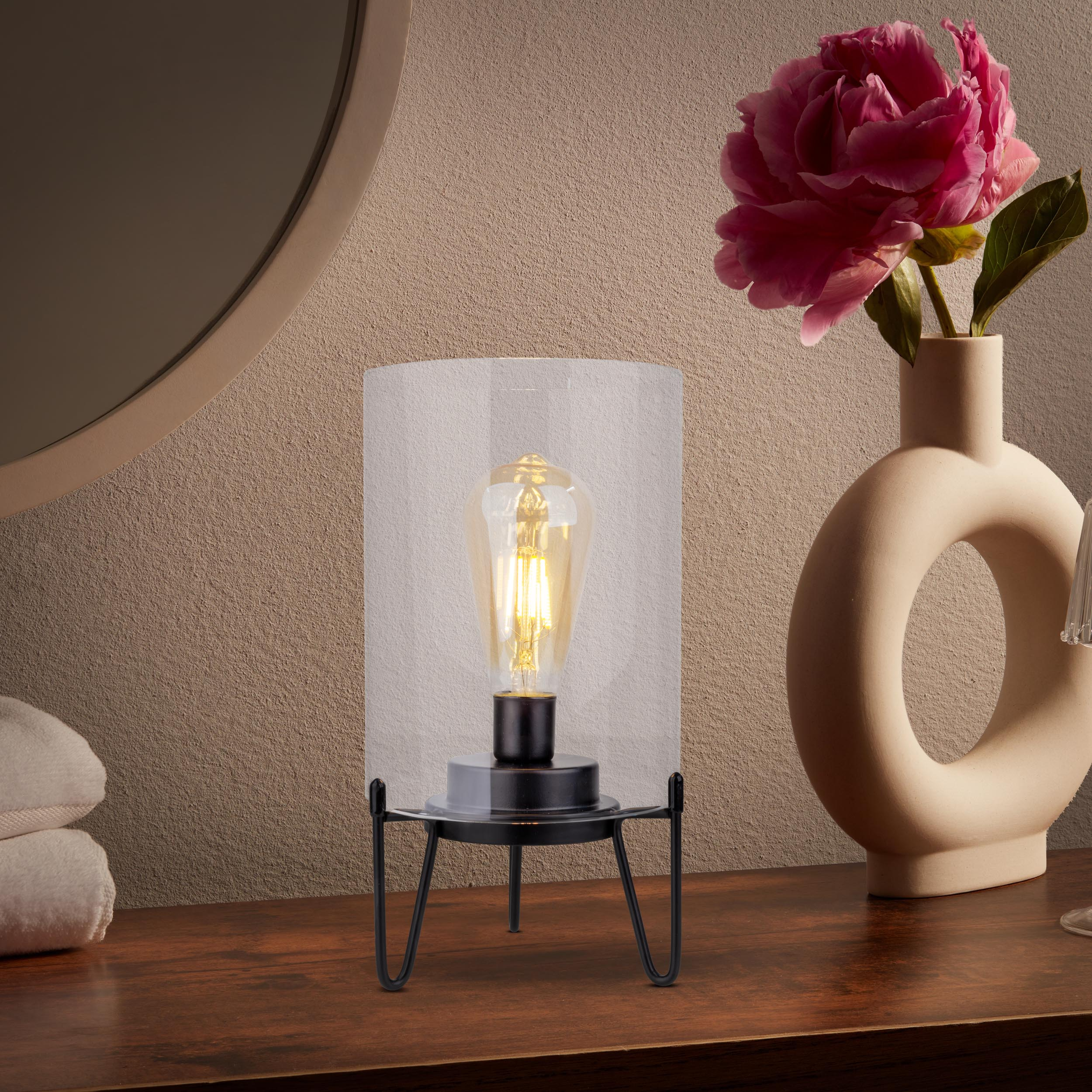 Eine Lampe mit Glühbirne in einem Glaszylinder steht auf Holz neben einer Vase mit rosa Blume.