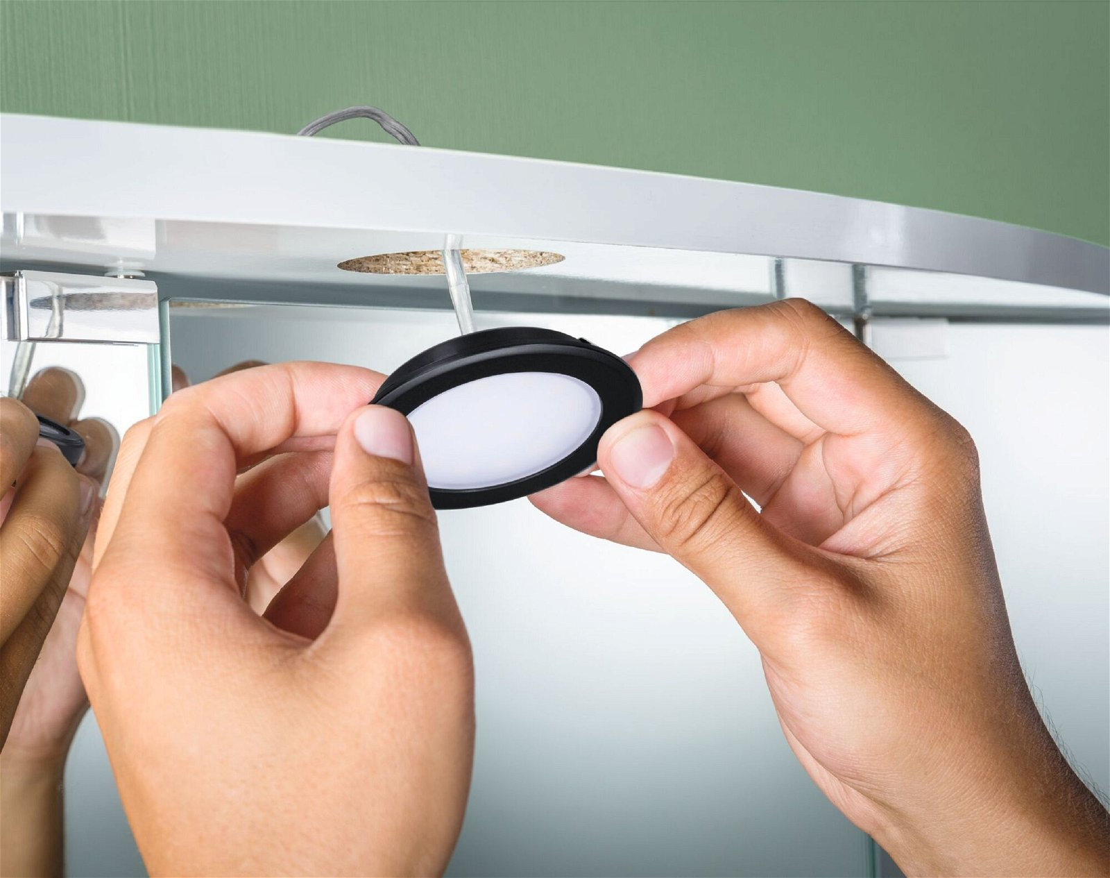 Ręce montujące okrągłą, czarną lampę LED w białej szafce.