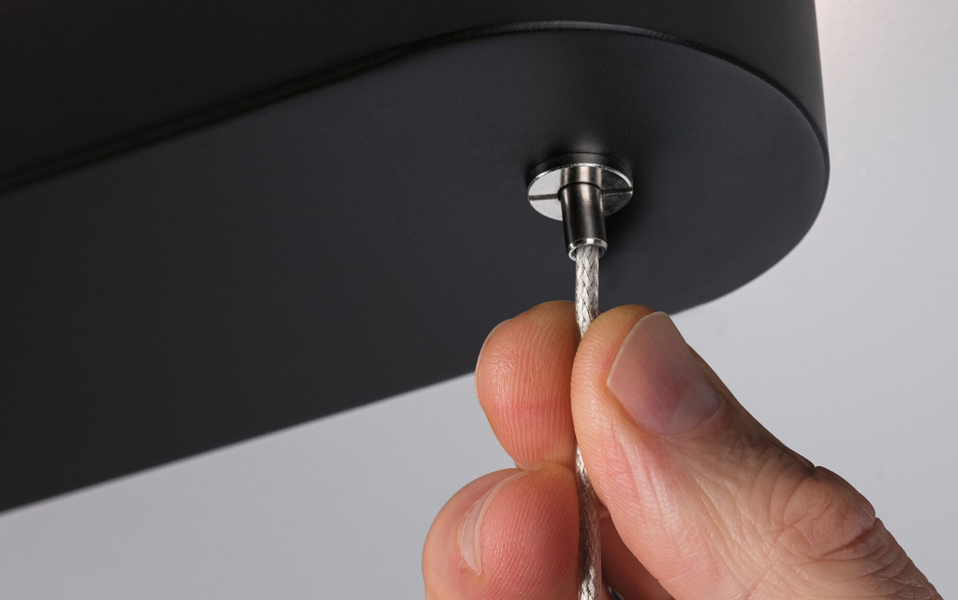 Ręka wkręcająca mały kabel do czarnej lampy sufitowej na jasnym tle.