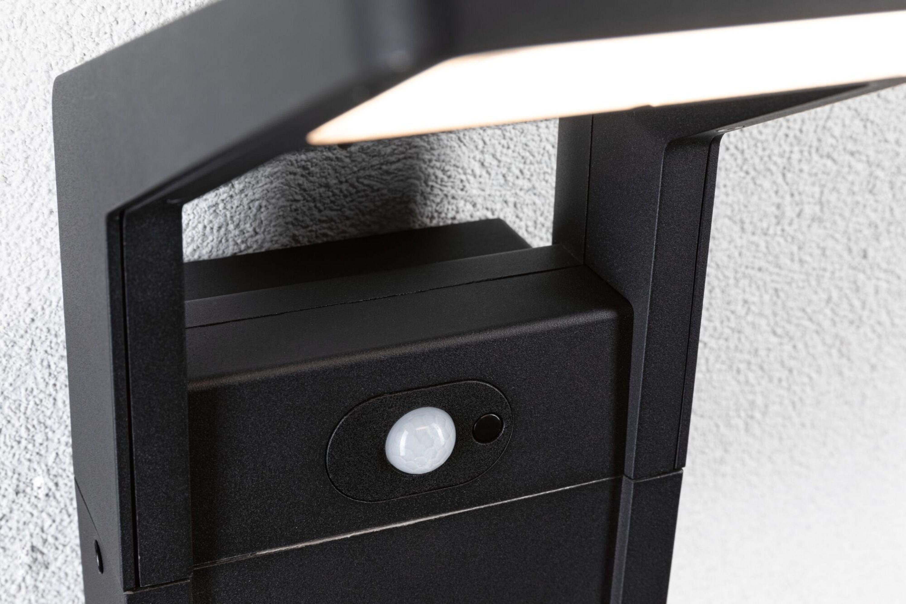 Czarna oprawa oświetleniowa zewnętrzna z czujnikiem. Światło jest włączone i oświetla.