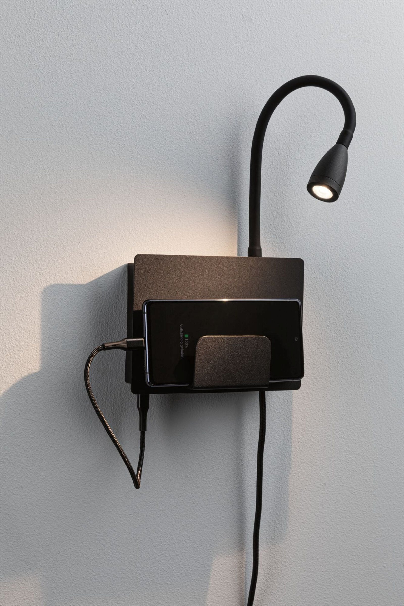 Czarna ładowarka naścienna z lampą z elastycznym ramieniem, telefon ładujący się, na białej ścianie.