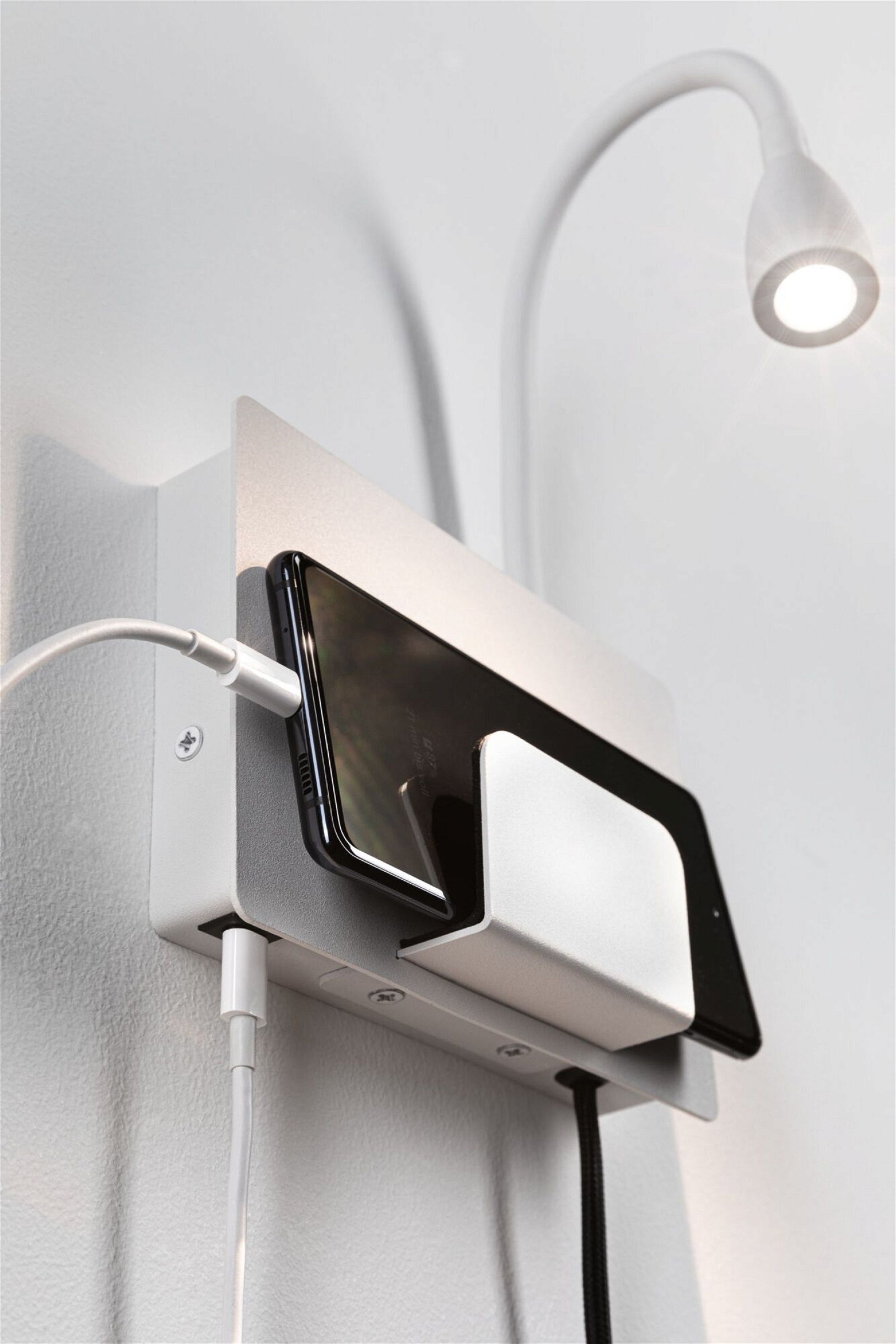 Biała, montowana na ścianie stacja ładująca z czarnym telefonem i tabletem. Mała lampa nad nimi.