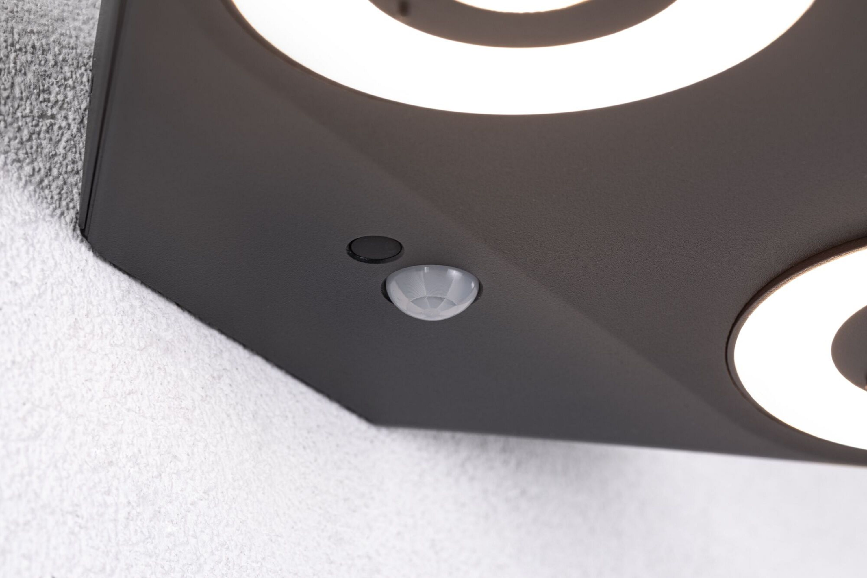 Zbliżenie czujnika PIR na ciemnej powierzchni. Tło: biała, teksturowana ściana.