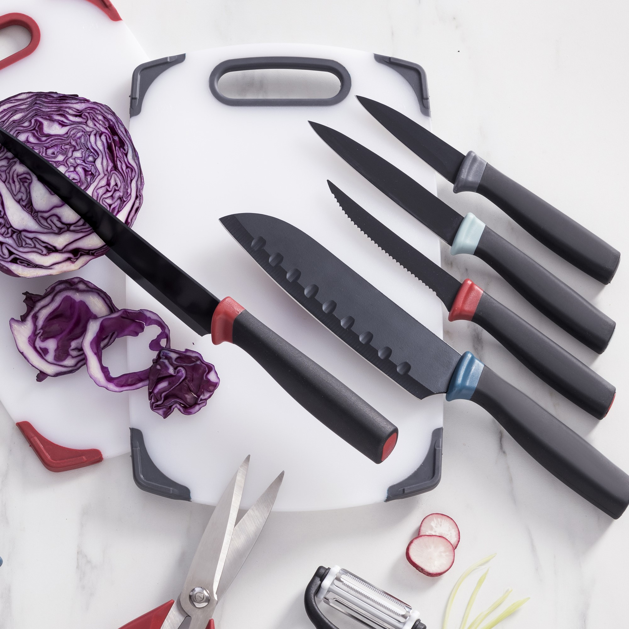 Un juego de cuchillos, tablas de cortar y una mitad de repollo morado en una superficie blanca.