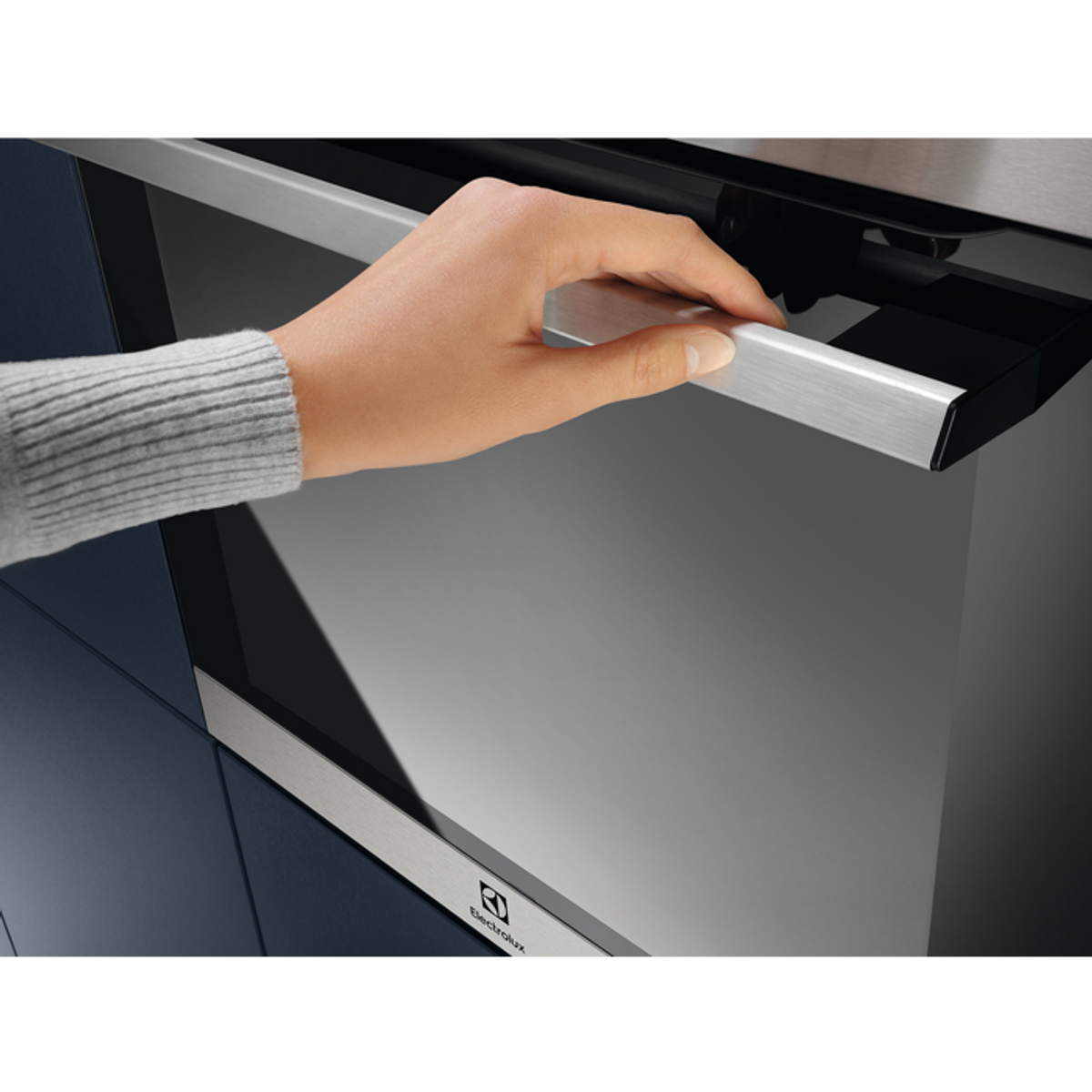 Ręka otwiera piekarnik. Drzwi piekarnika mają srebrną rączkę. Tło przedstawia powierzchnię piekarnika i logo Electrolux.