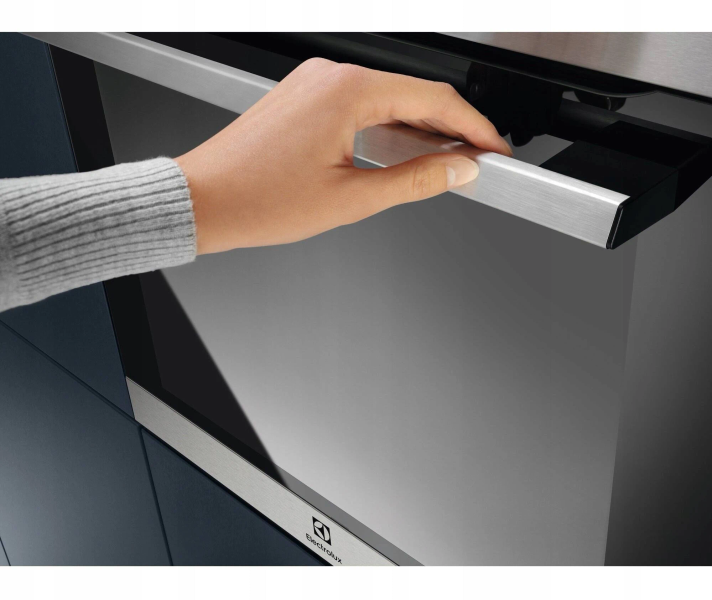 Ręka otwiera drzwi piekarnika. Piekarnik jest czarny z metalowym uchwytem i ramą. Ściana jest ciemnoniebieska.