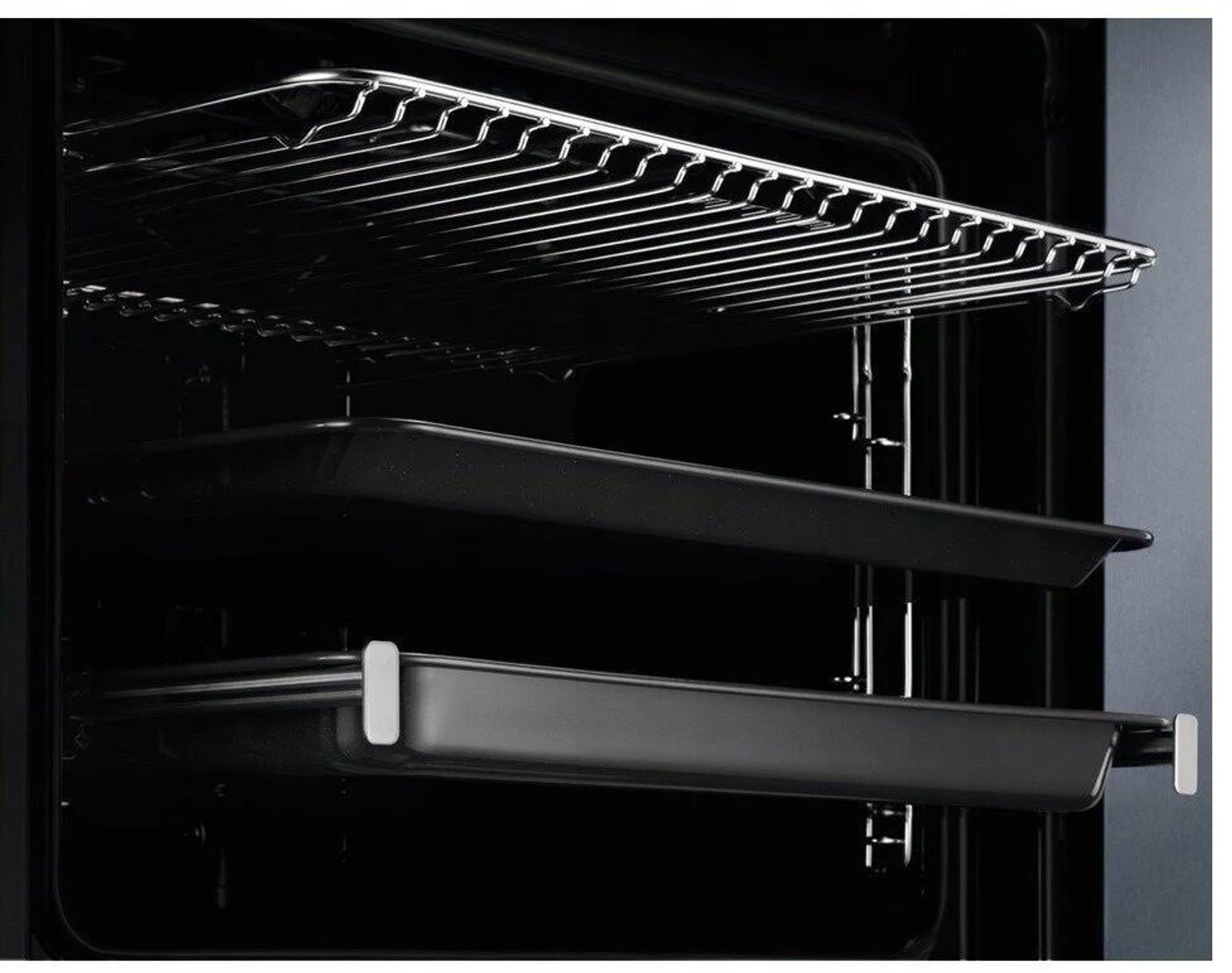 Wewnątrz piekarnika widoczny jest druciany ruszt i dwie czarne tace. Wnętrze piekarnika jest ciemne. Widoczna jest rama drzwi.
