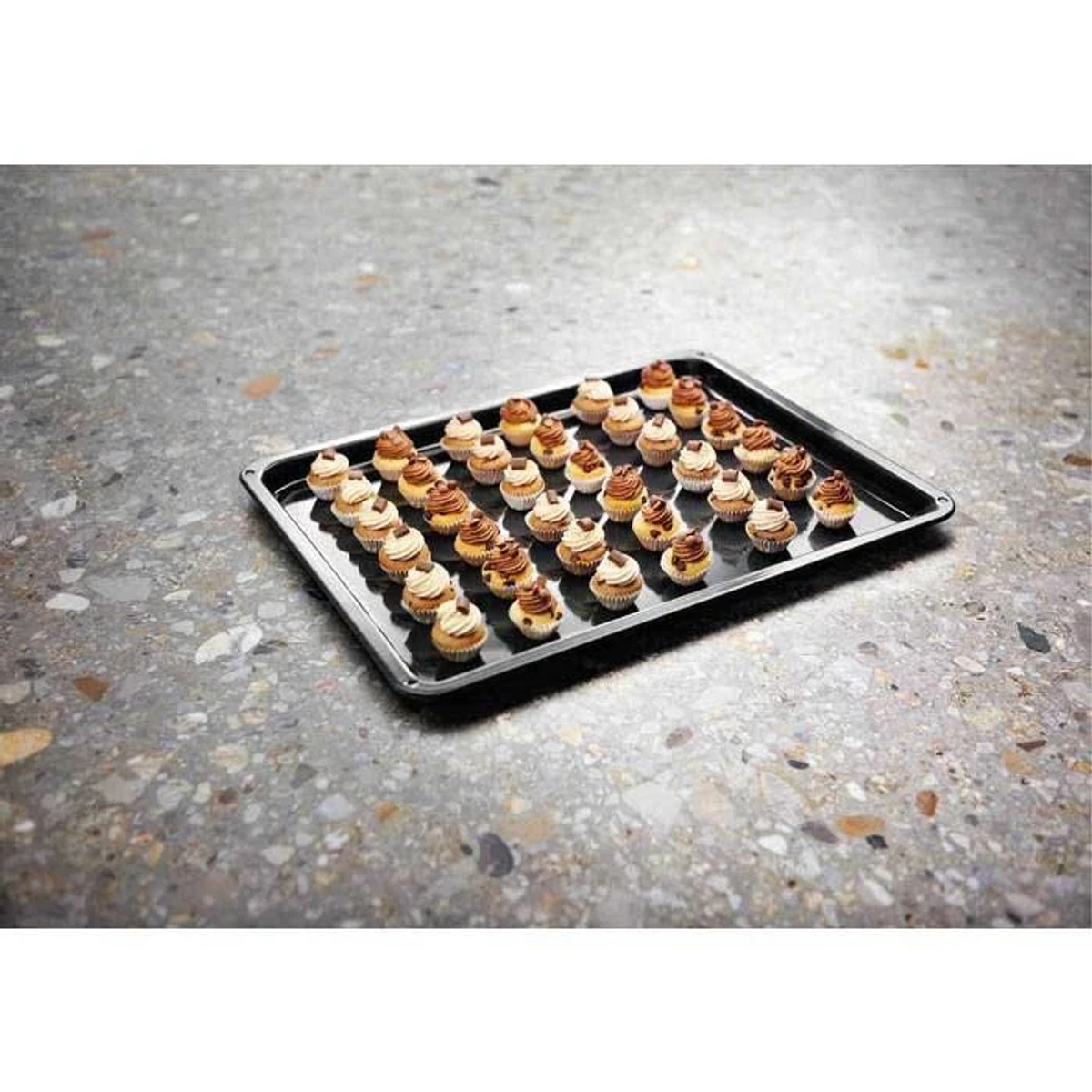 Blacha do pieczenia zawiera wiele mini babeczek. Mają białe i brązowe polewy. Taca stoi na nakrapianej szarej powierzchni.