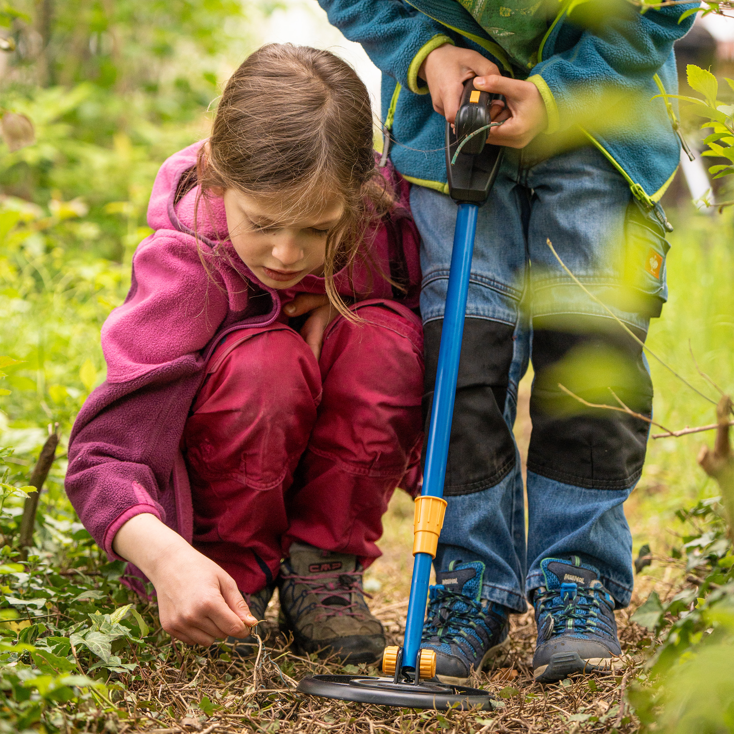 Dos niños exploran un bosque con un detector de metales. Uno está arrodillado, mirando algo.