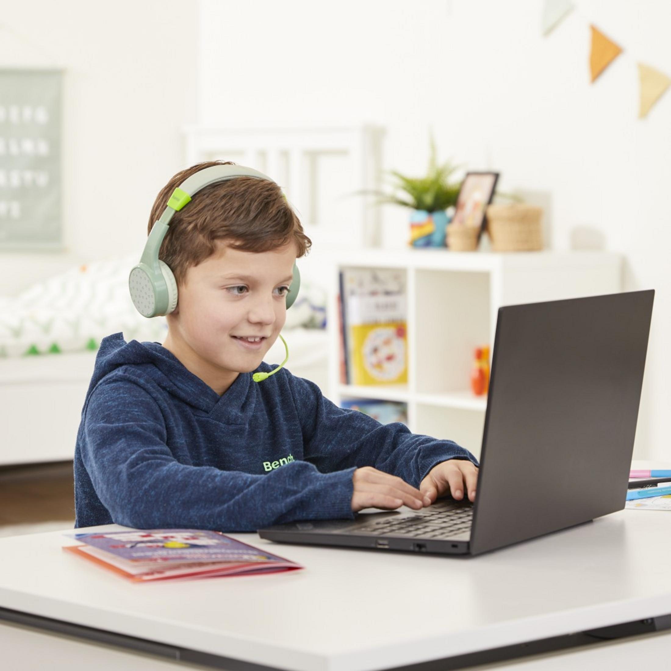 Junge mit Kopfhörern benutzt Laptop an weißem Schreibtisch. Karte und Bücher vorhanden.