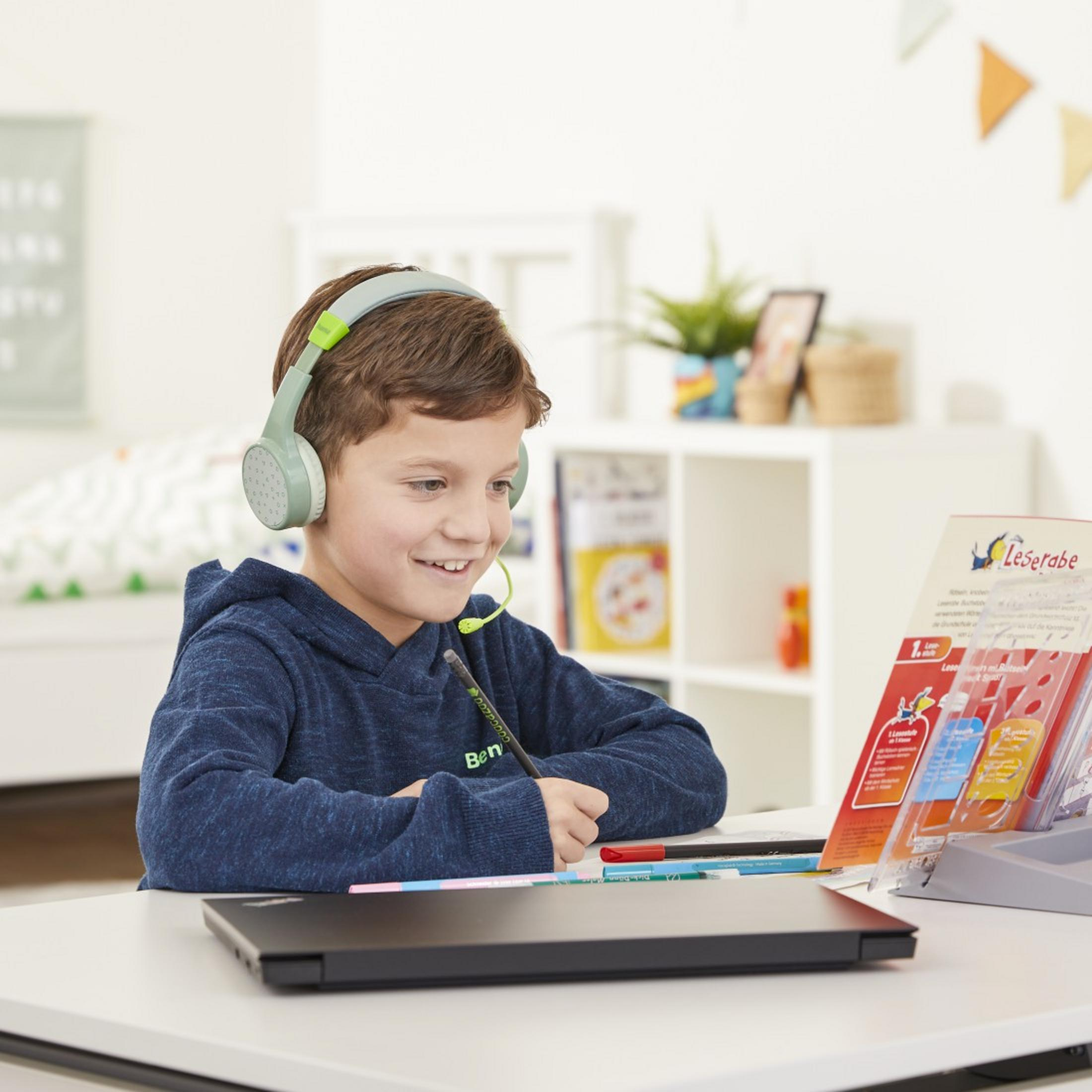 Junge mit Headset, schreibt auf Papier, Laptop auf dem Schreibtisch. Buchständer rechts.