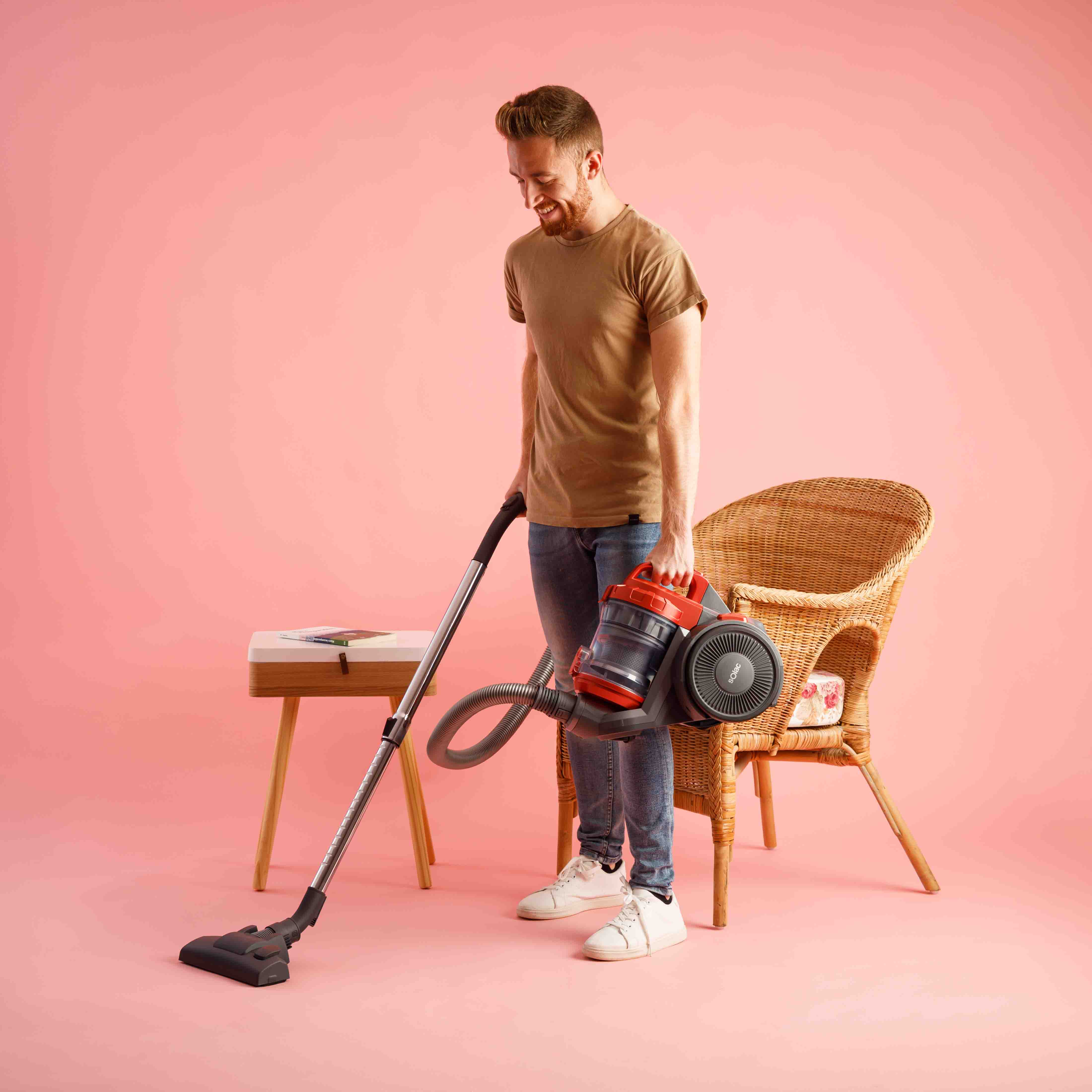 Mężczyzna odkurza pokój. Trzyma odkurzacz i wąż, uśmiechając się. W scenie znajduje się wiklinowy fotel i stół.