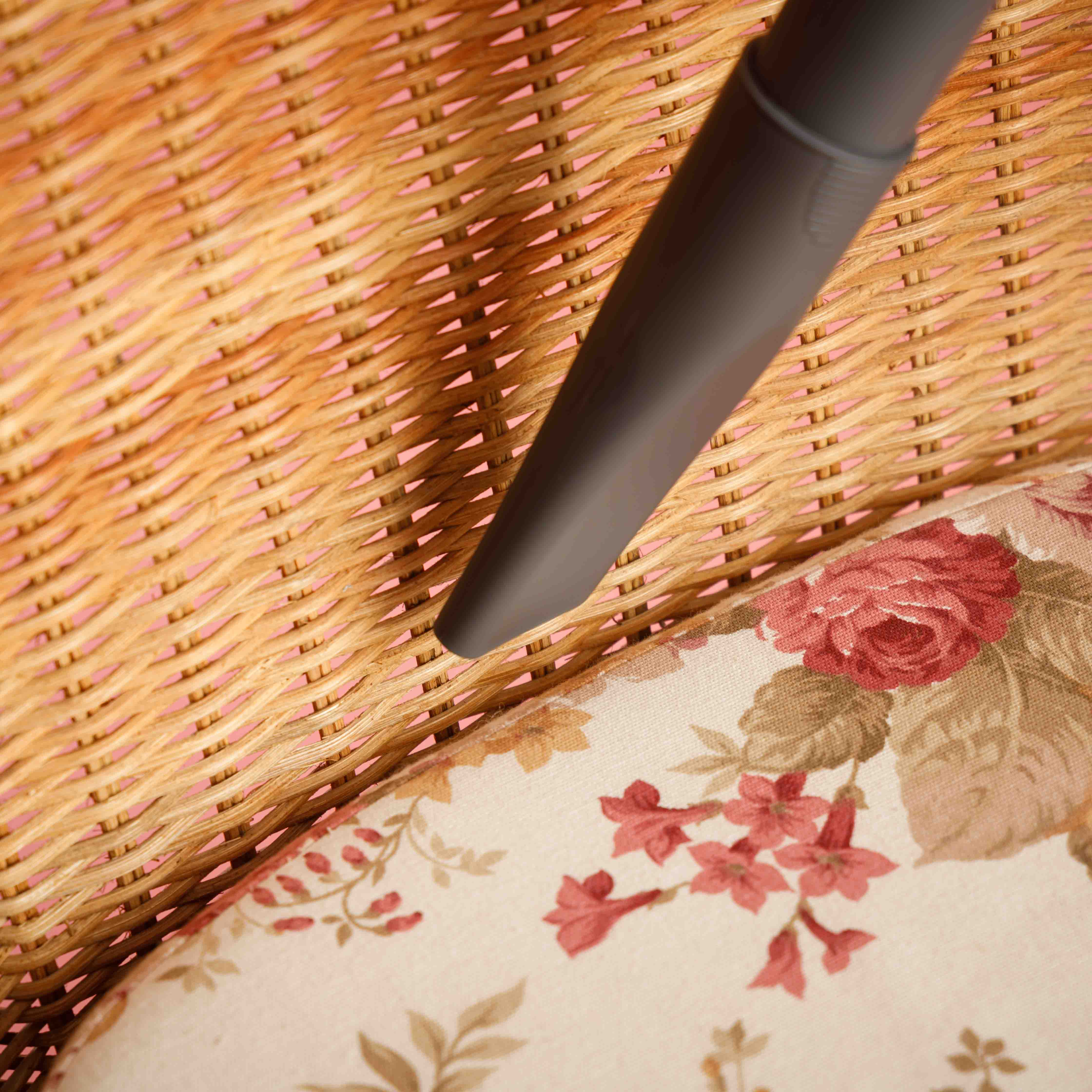 Cabezal de aspiradora limpiando una silla de mimbre con cojín floral. Fondo rosa.