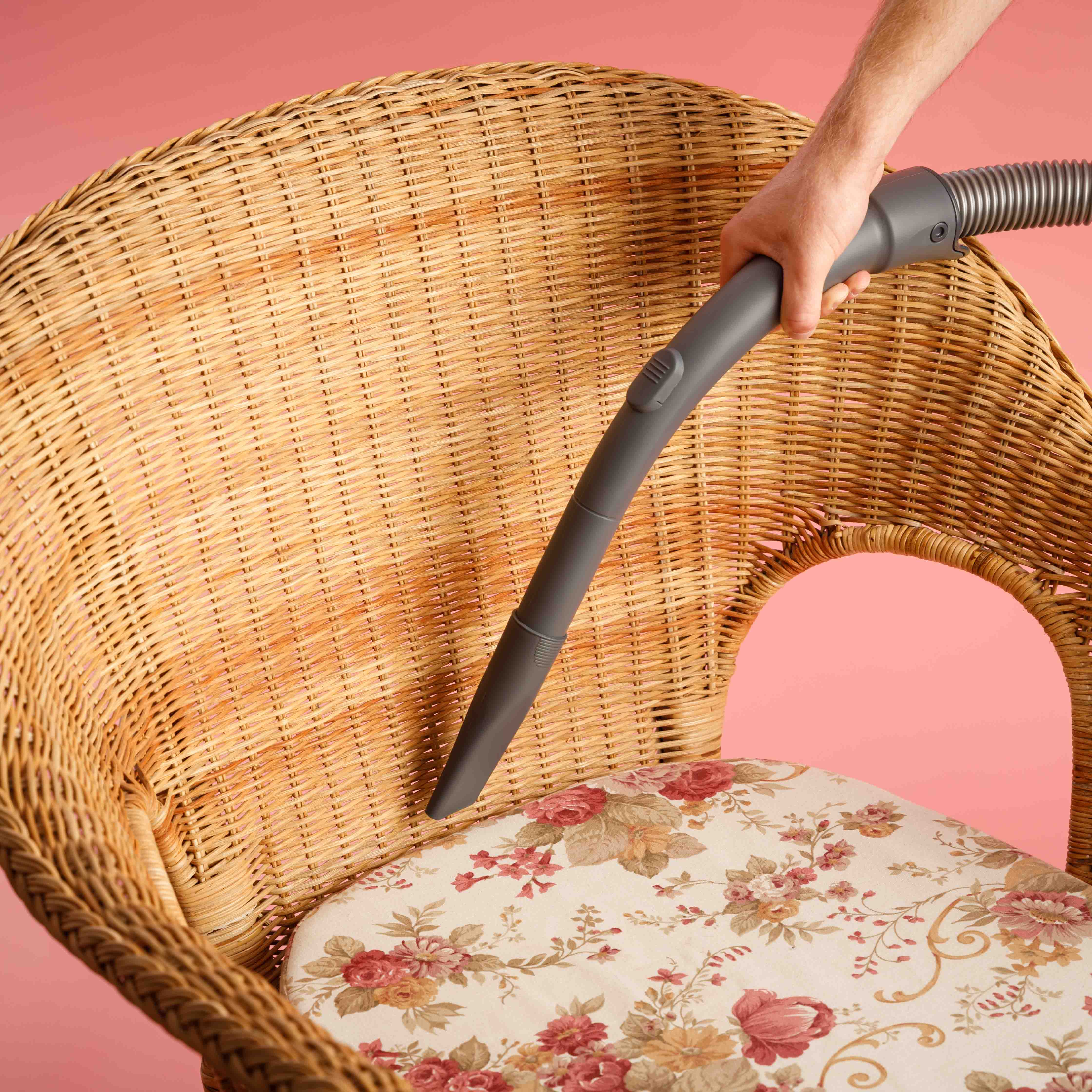 Una mano usando una boquilla de aspiradora en una silla de mimbre con cojín floral.