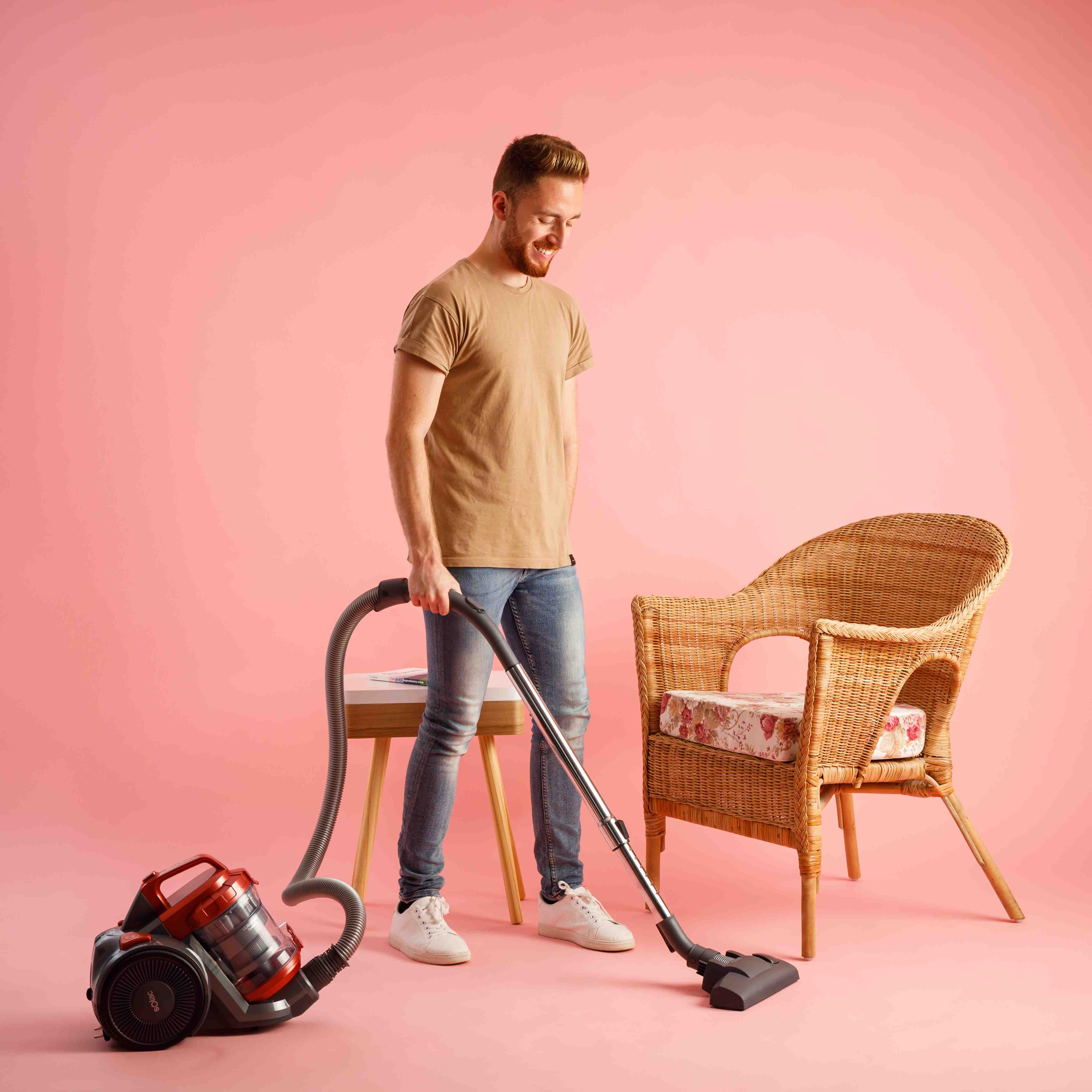 Hombre aspirando habitación con pared rosa, cerca de silla mimbre y mesa pequeña.