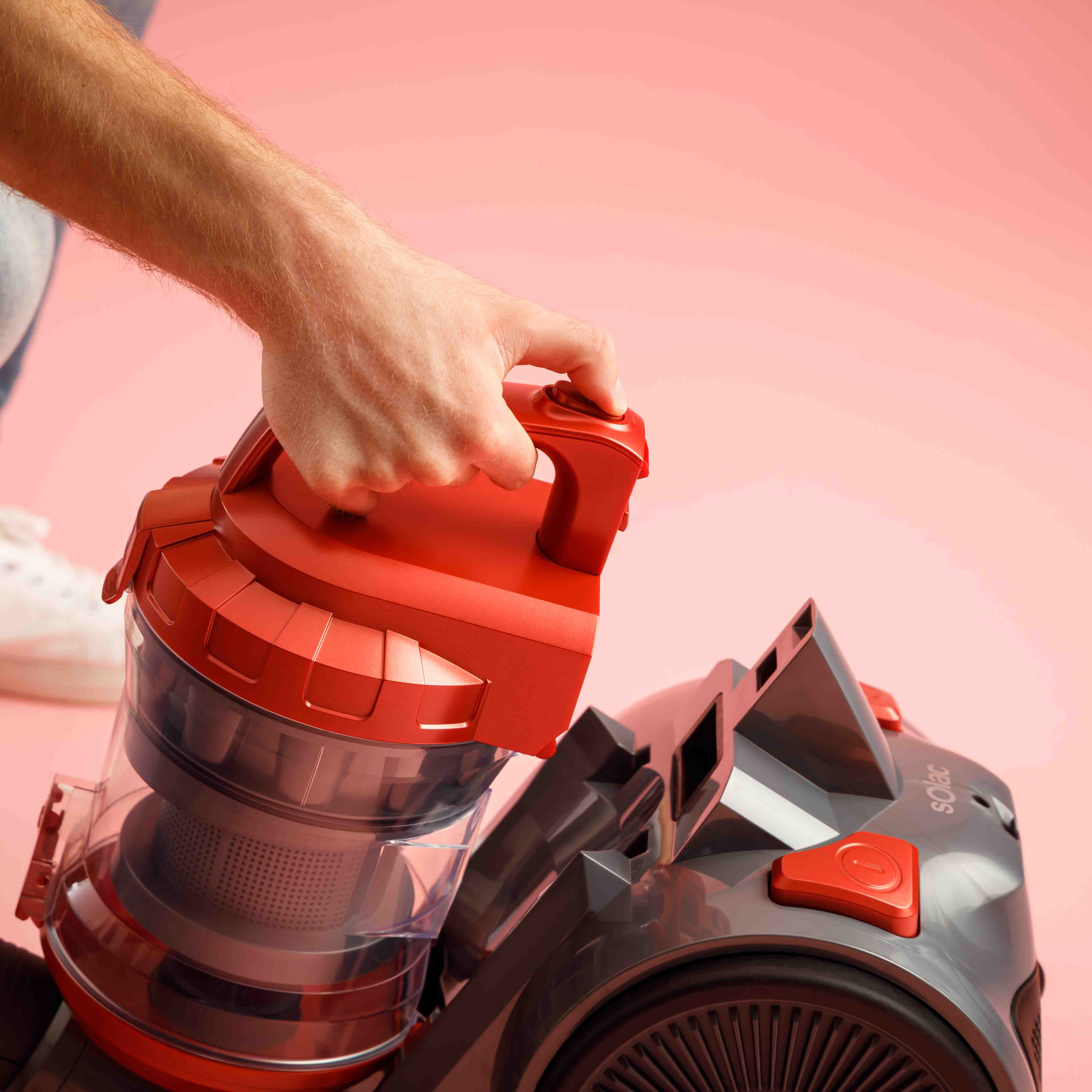Dłoń trzymająca czerwony uchwyt odkurzacza, widoczna część pojemnika.