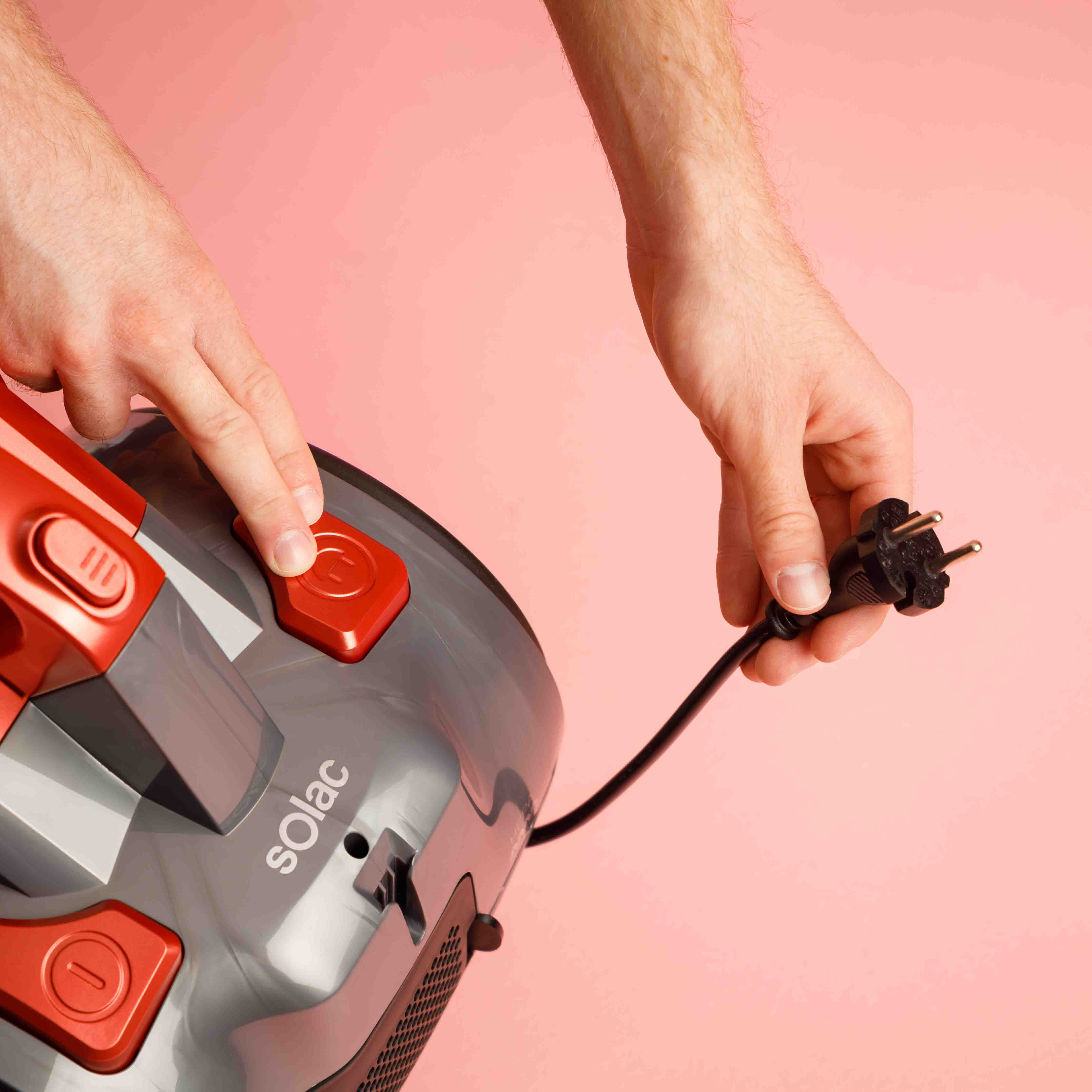 Ręce podłączające przewód zasilający do szaro-czerwonego odkurzacza, na różowym tle.