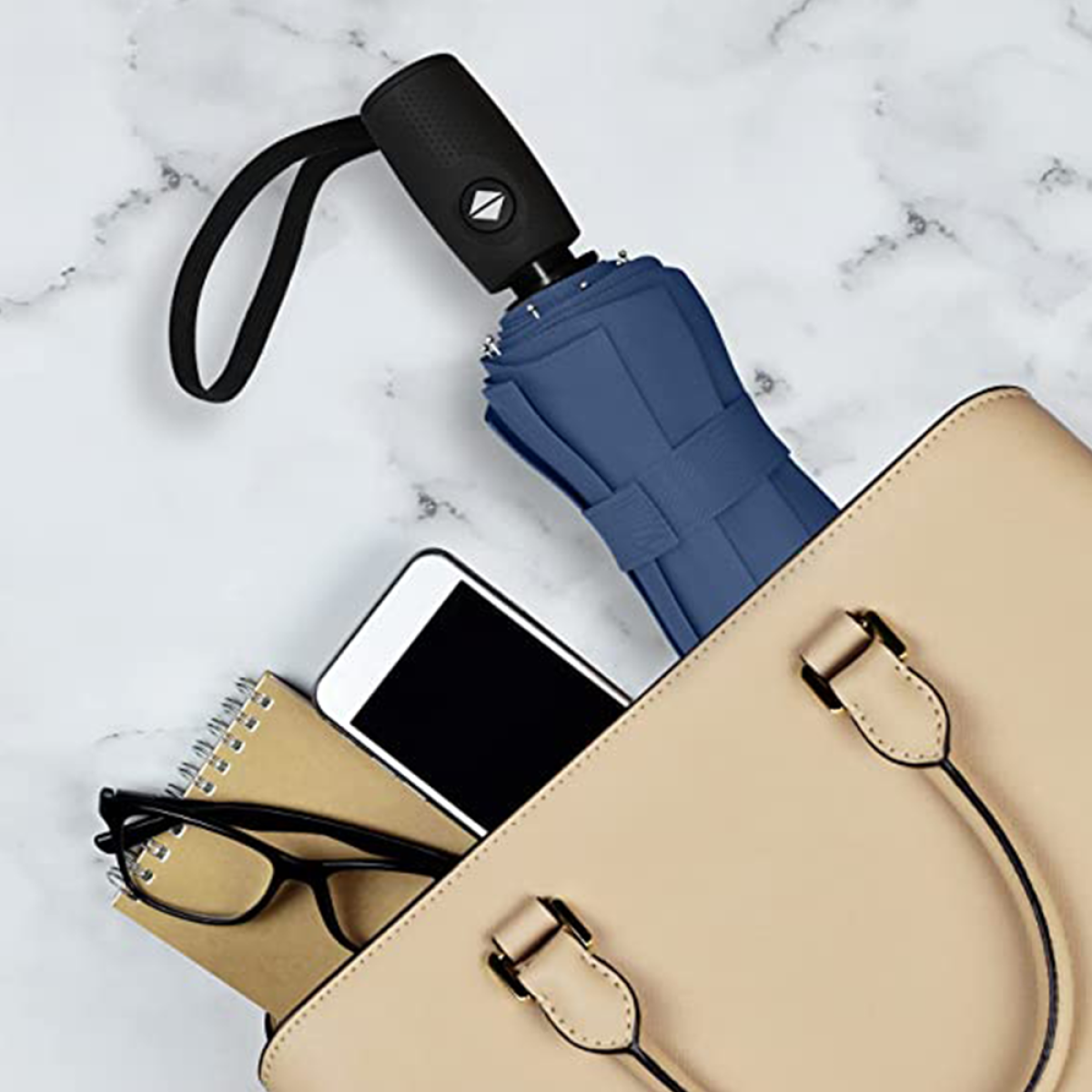 Ein blauer geschlossener Regenschirm in einer beigen Handtasche, mit Telefon, Brille.