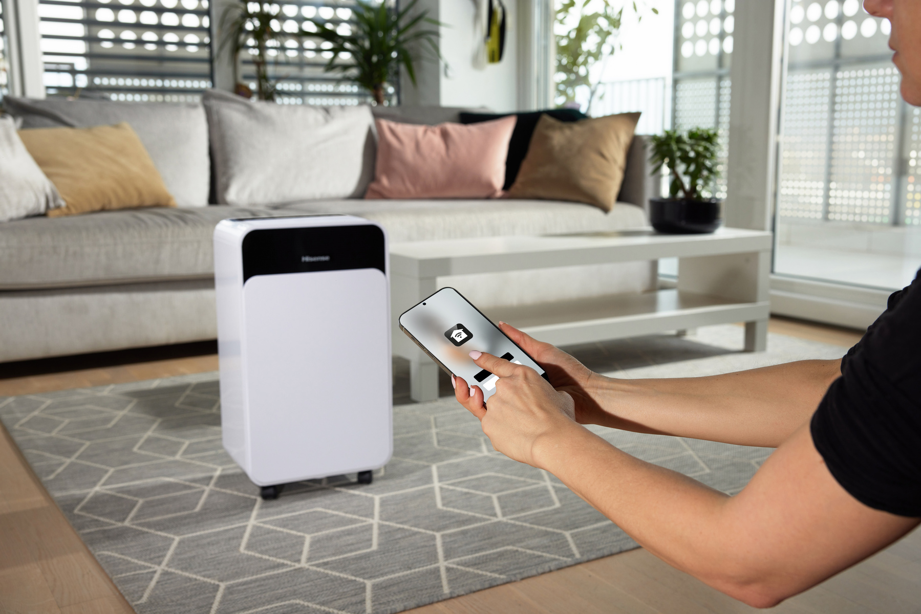 Osoba trzymająca telefon kontrolująca biały oczyszczacz powietrza w salonie.