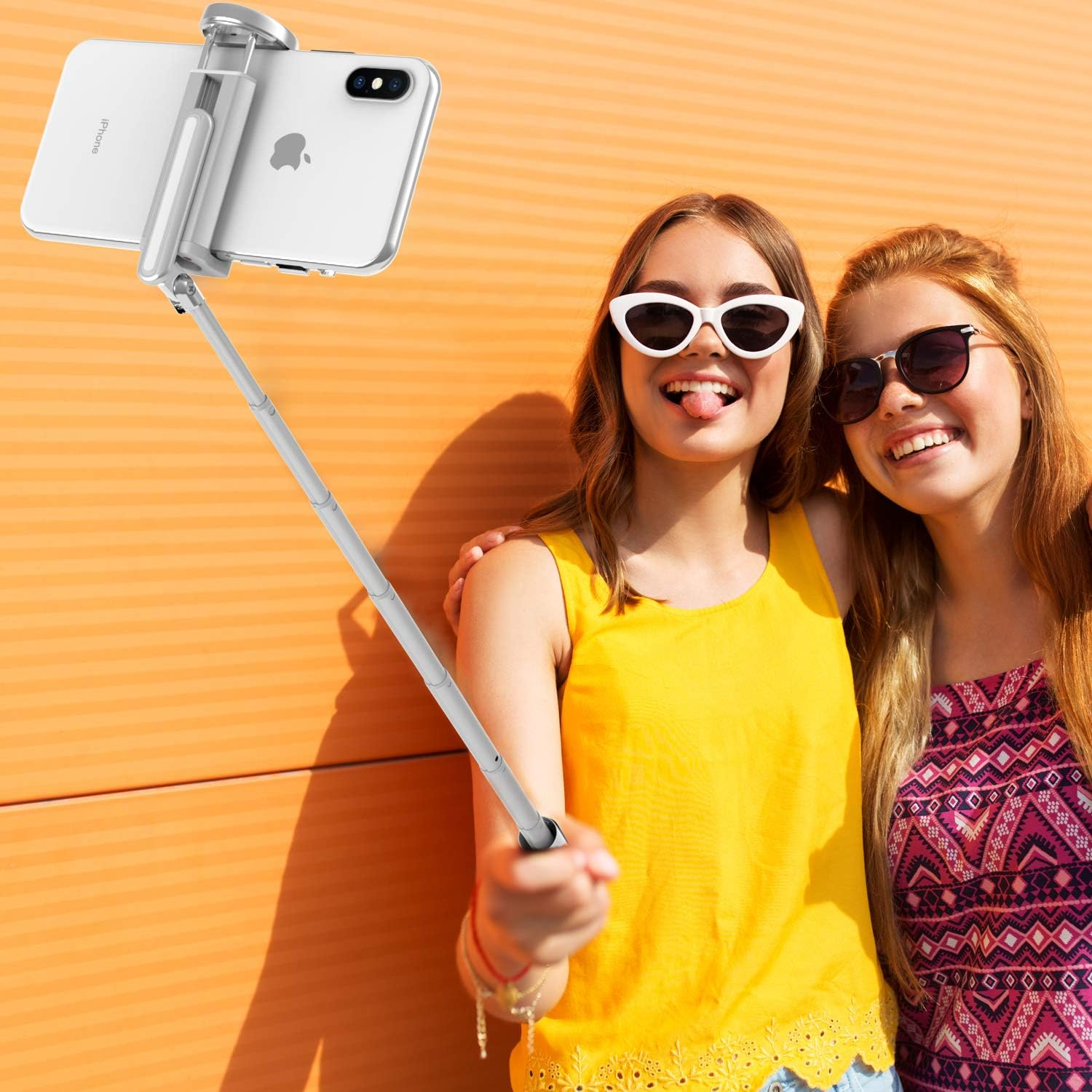 Zwei junge Frauen mit Selfie-Stick. Eine streckt die Zunge raus, die andere lächelt. Oranger Hintergrund.