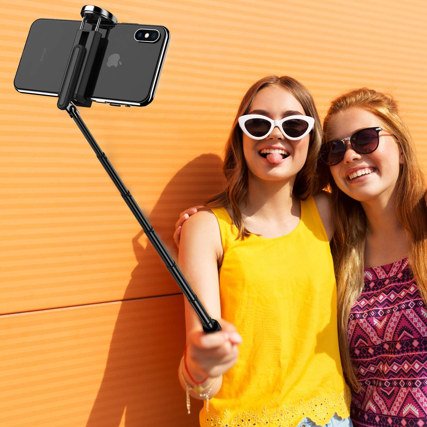 Zwei Mädchen mit Sonnenbrillen machen ein Selfie mit einem Selfie-Stick vor orangefarbenem Hintergrund.