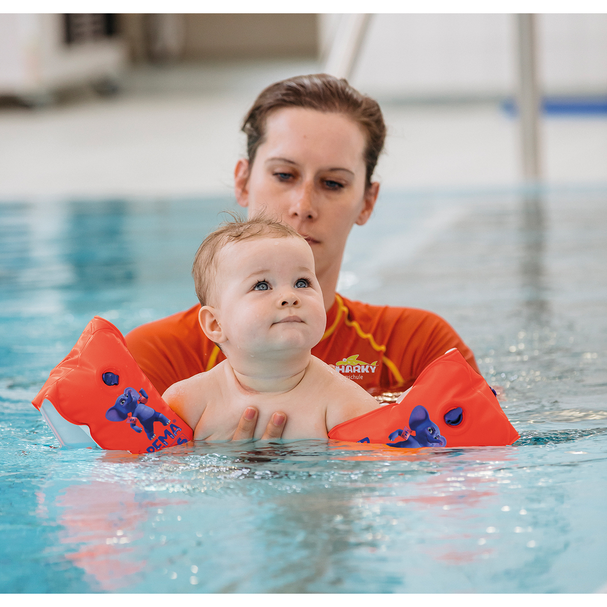 Ein Baby im Wasser mit Armschwimmflügeln, das von einem Erwachsenen gehalten wird.
