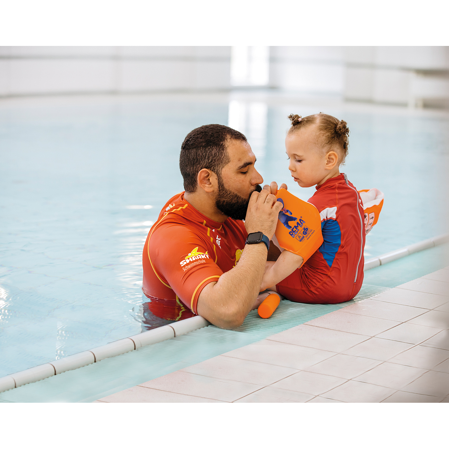 Ein Mann und ein Kleinkind in einem Schwimmbecken. Der Mann hilft dem Kind mit einer Schwimmhilfe.