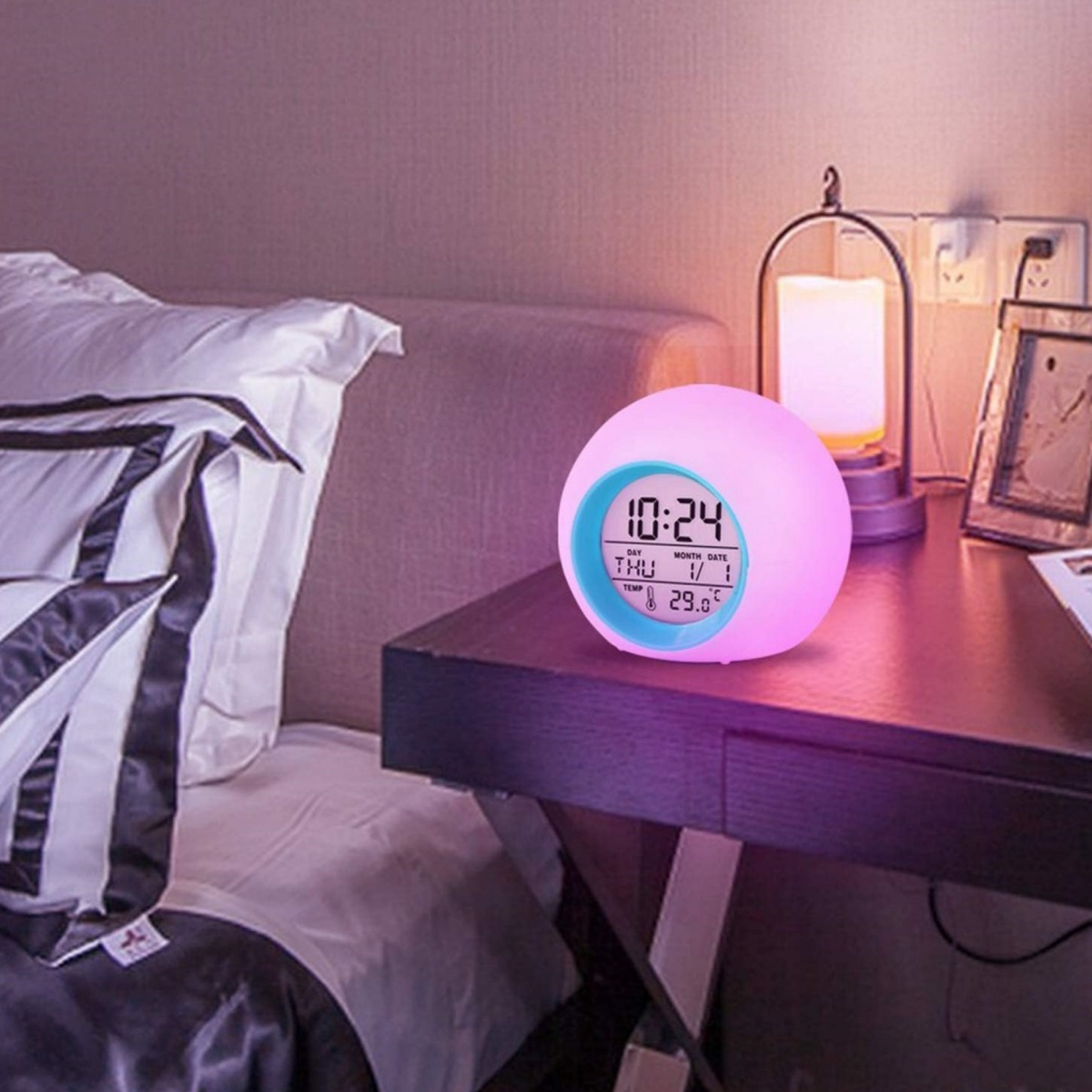 Eine leuchtende Uhr zeigt Zeit und Datum an und steht auf einem dunklen Tisch. Im Hintergrund sind ein Bett und eine Lampe.