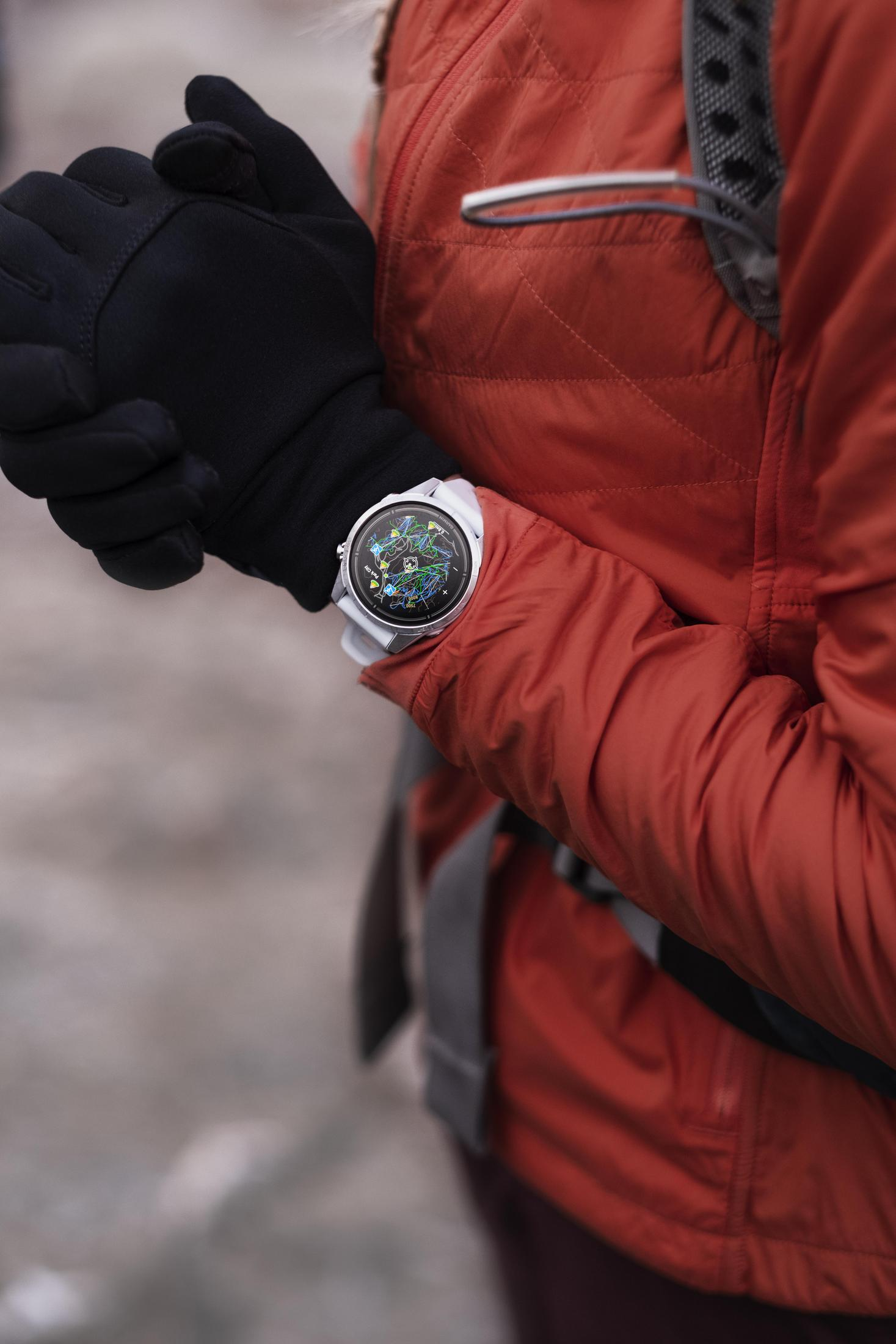 Osoba nosząca zegarek na nadgarstku i czarne rękawiczki. Zegarek jest biały z kolorowym wyświetlaczem.