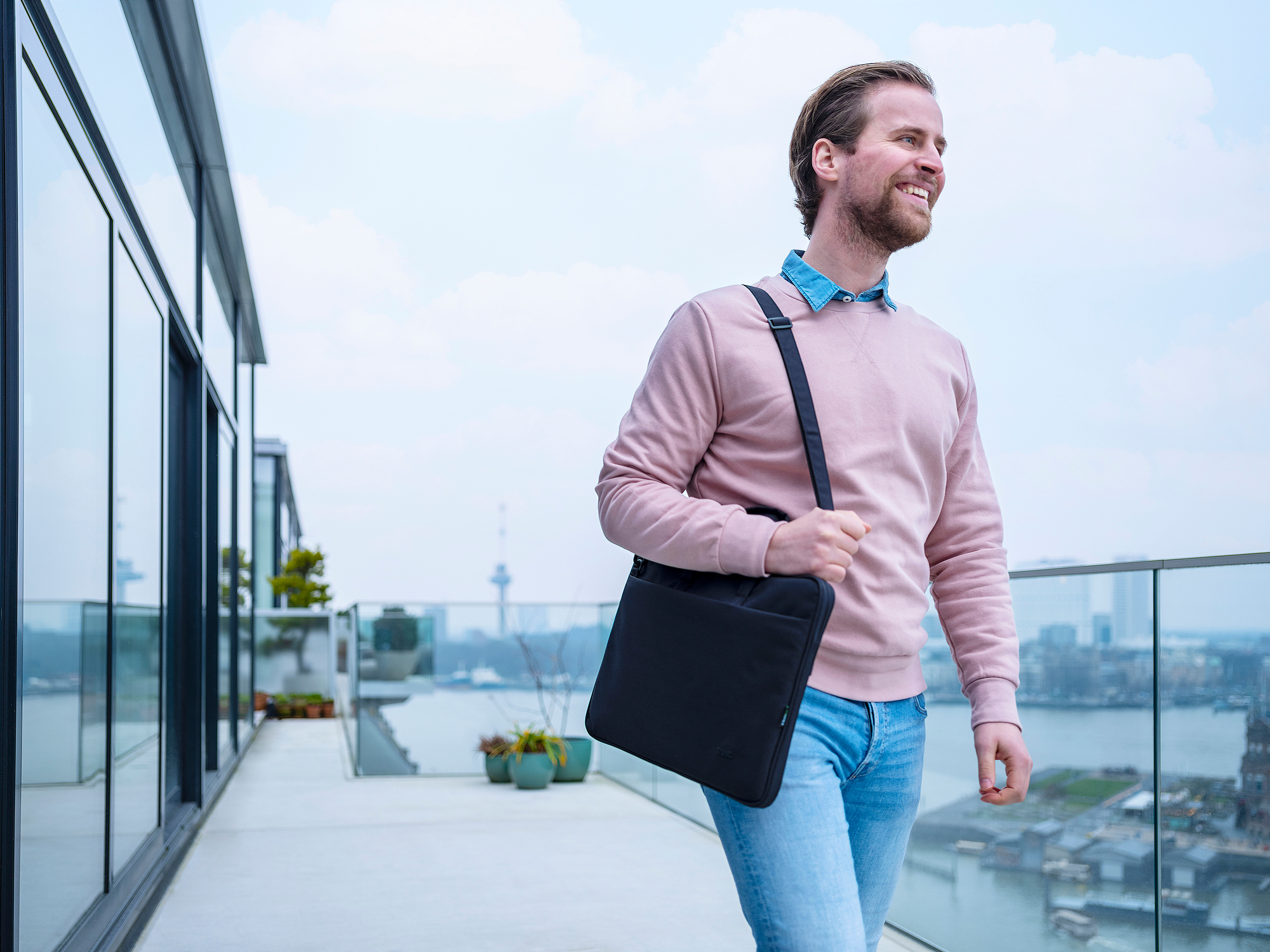 A man with a pink sweater and jeans carrying a black laptop bag. He is smiling.