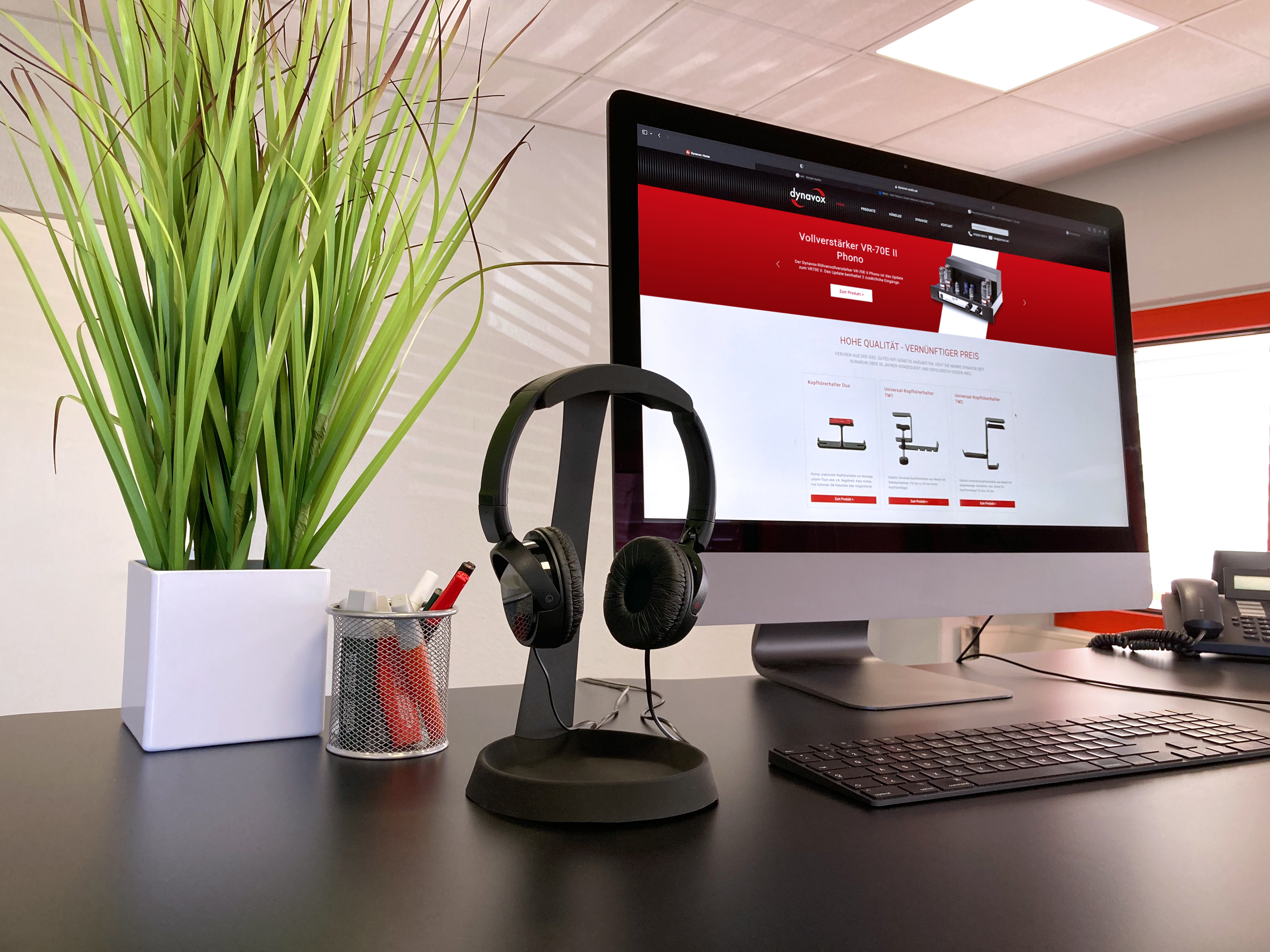 Bürotisch mit Bildschirm, Kopfhörern und Pflanze. Webseite wird auf dem Bildschirm angezeigt.
