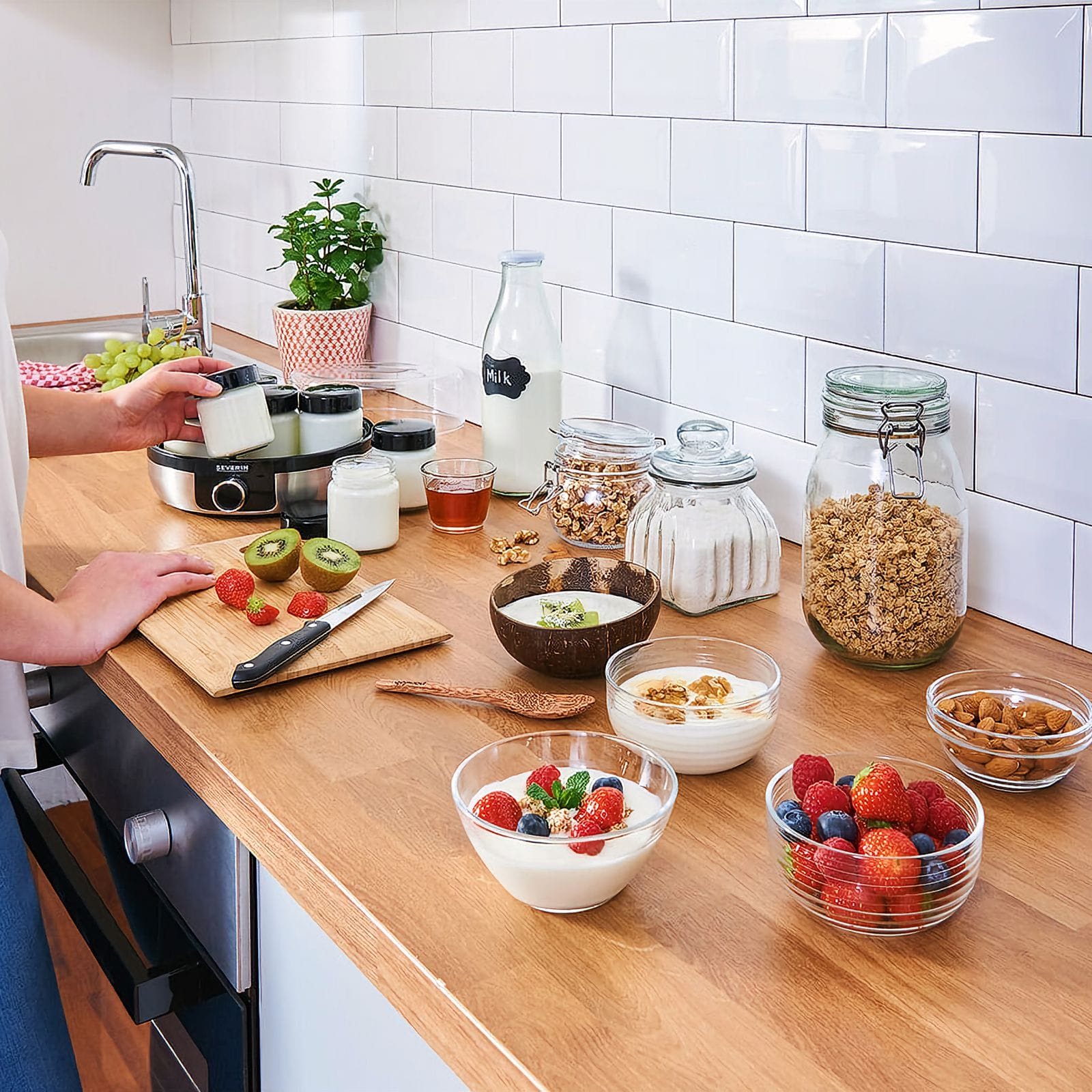 Una encimera de cocina con elementos de desayuno: yogur con bayas, granola, fruta, leche y una yogurtera.