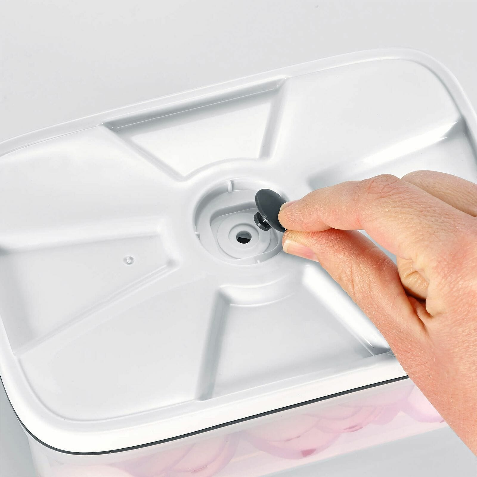 Coperchio bianco del contenitore con una mano che preme una valvola nera. Cipolle rosa all'interno. Lo sfondo è bianco.